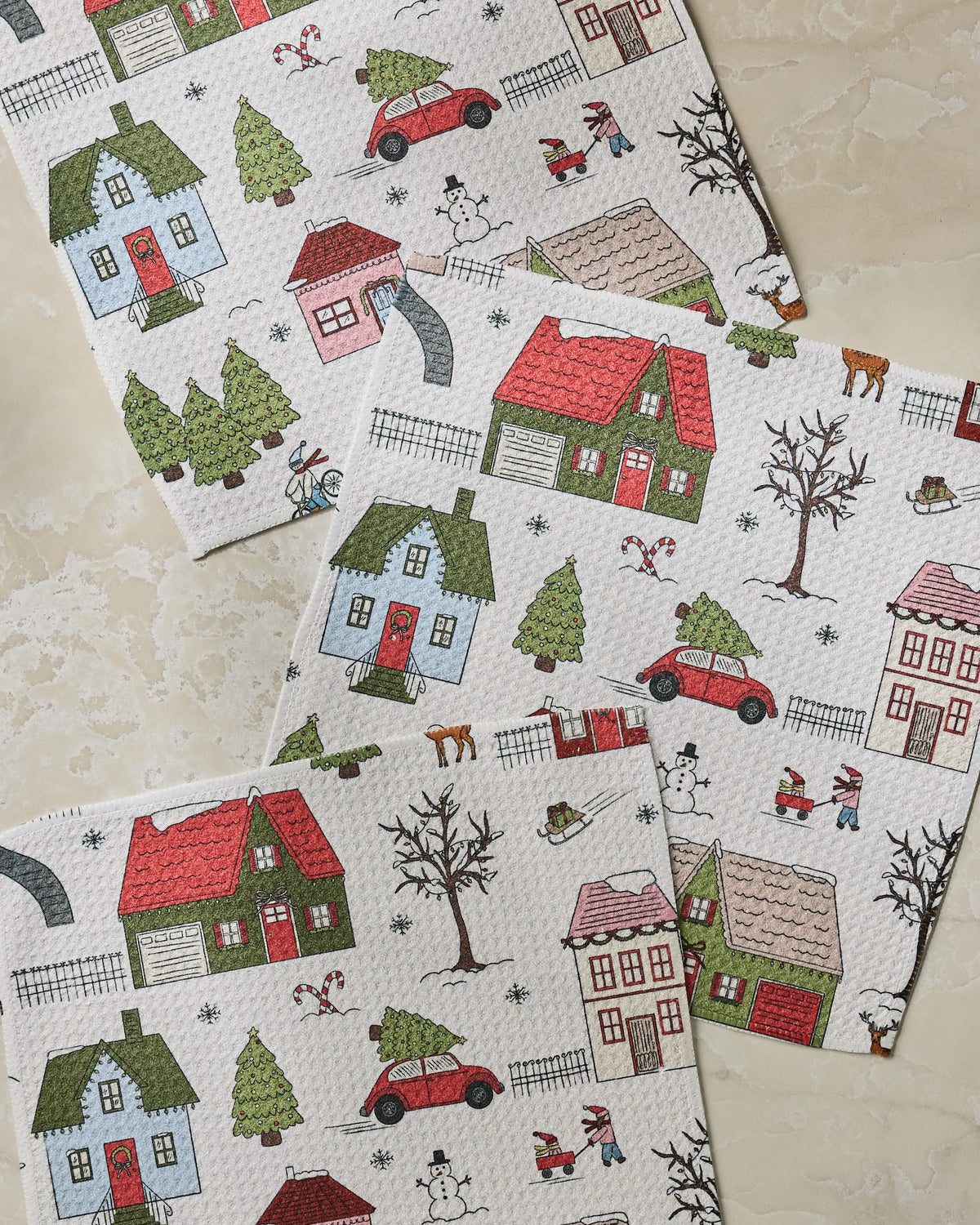 Geometry’s Winter Village paper towels, crafted from recycled materials, display a delightful pattern of houses, cars, trees, snowmen, and children playing in the snow on a light background—beautifully arranged on a stone surface.