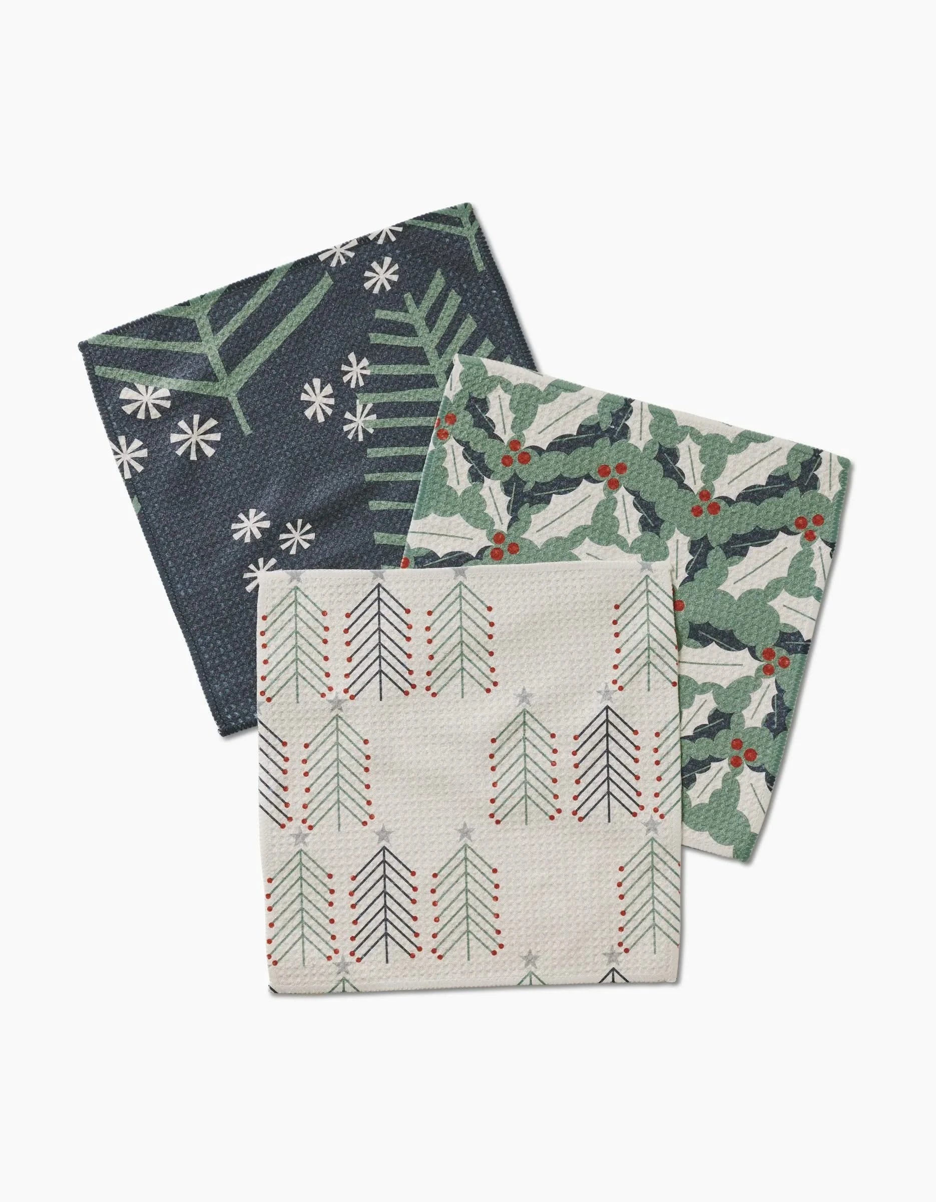 The Winter Wonderful napkin set by Lizzie Clark features three Geoweave fabric napkins: one with white snowflakes and green branches on dark blue, one with green holly and red berries, and one with stylized trees in red, green, and blue on beige.