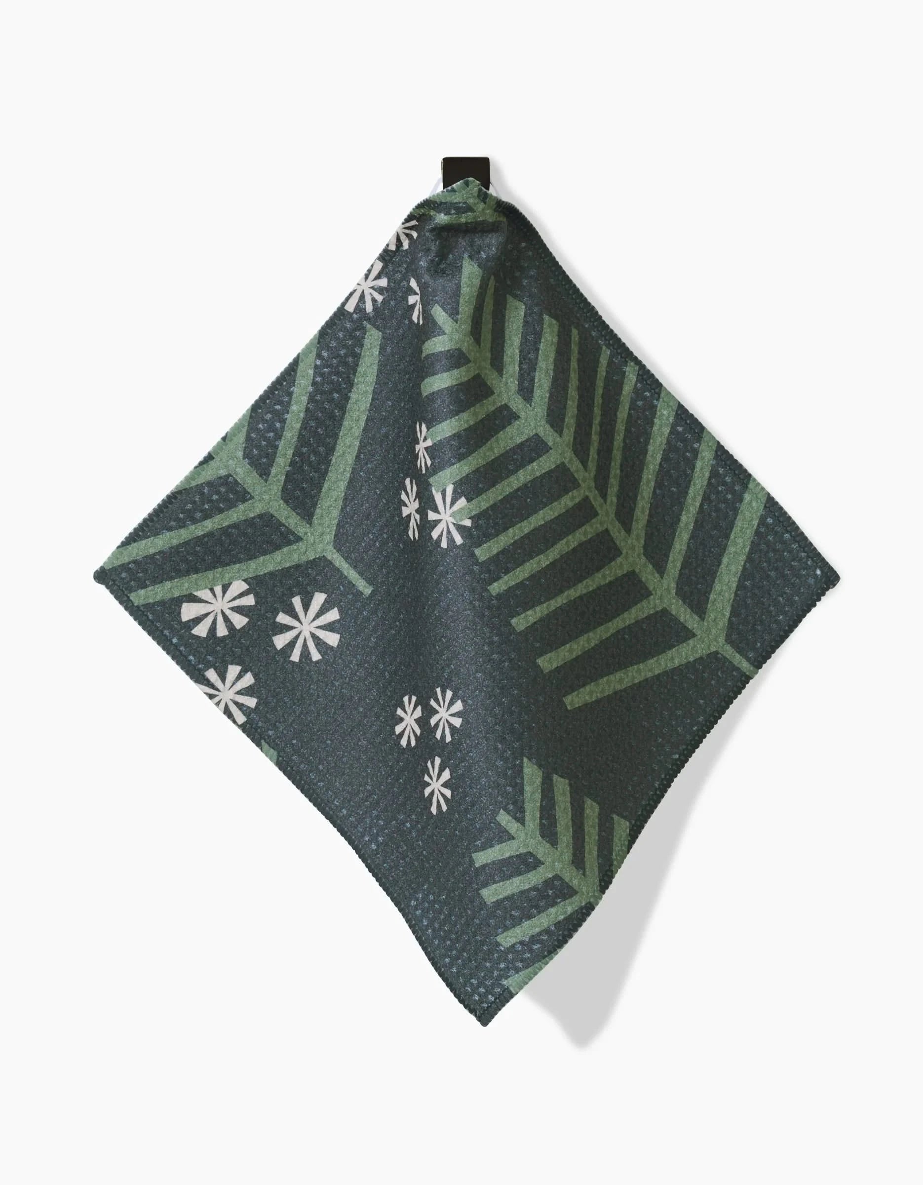 The Lizzie Clark Winter Wonderful napkin features dark green textured Geoweave fabric with light green leaf patterns and small white asterisk-like flowers. It hangs by a black loop against a white background and is made from recycled materials.