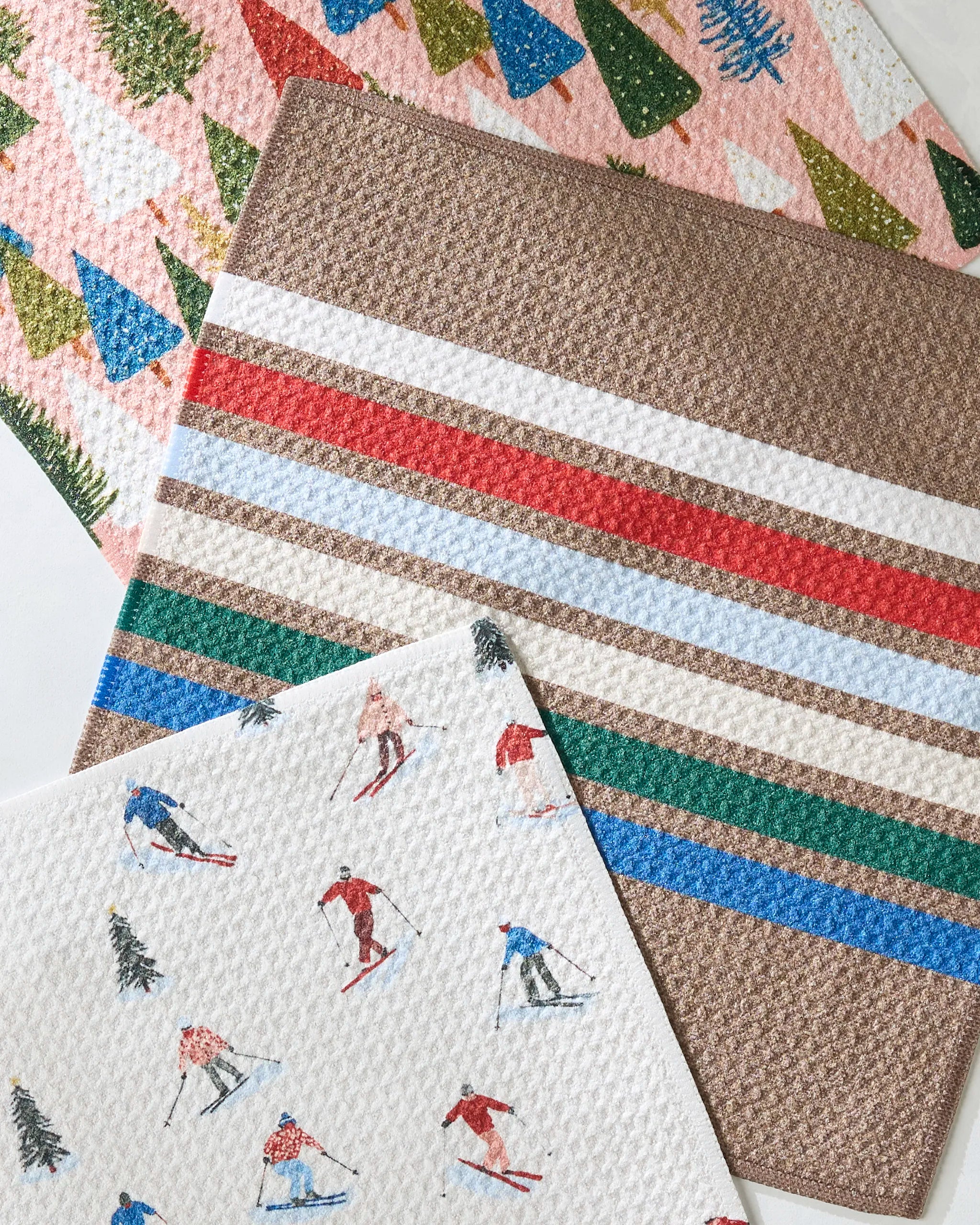 Three absorbent Geometry Winterhaus dishcloths overlap: one with colorful trees, one with horizontal brown and multicolored stripes, and one with skiers and trees—an eco-friendly recycled set for your kitchen.
