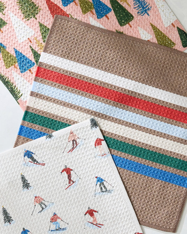 Three absorbent Geometry Winterhaus dishcloths overlap: one with colorful trees, one with horizontal brown and multicolored stripes, and one with skiers and trees—an eco-friendly recycled set for your kitchen.