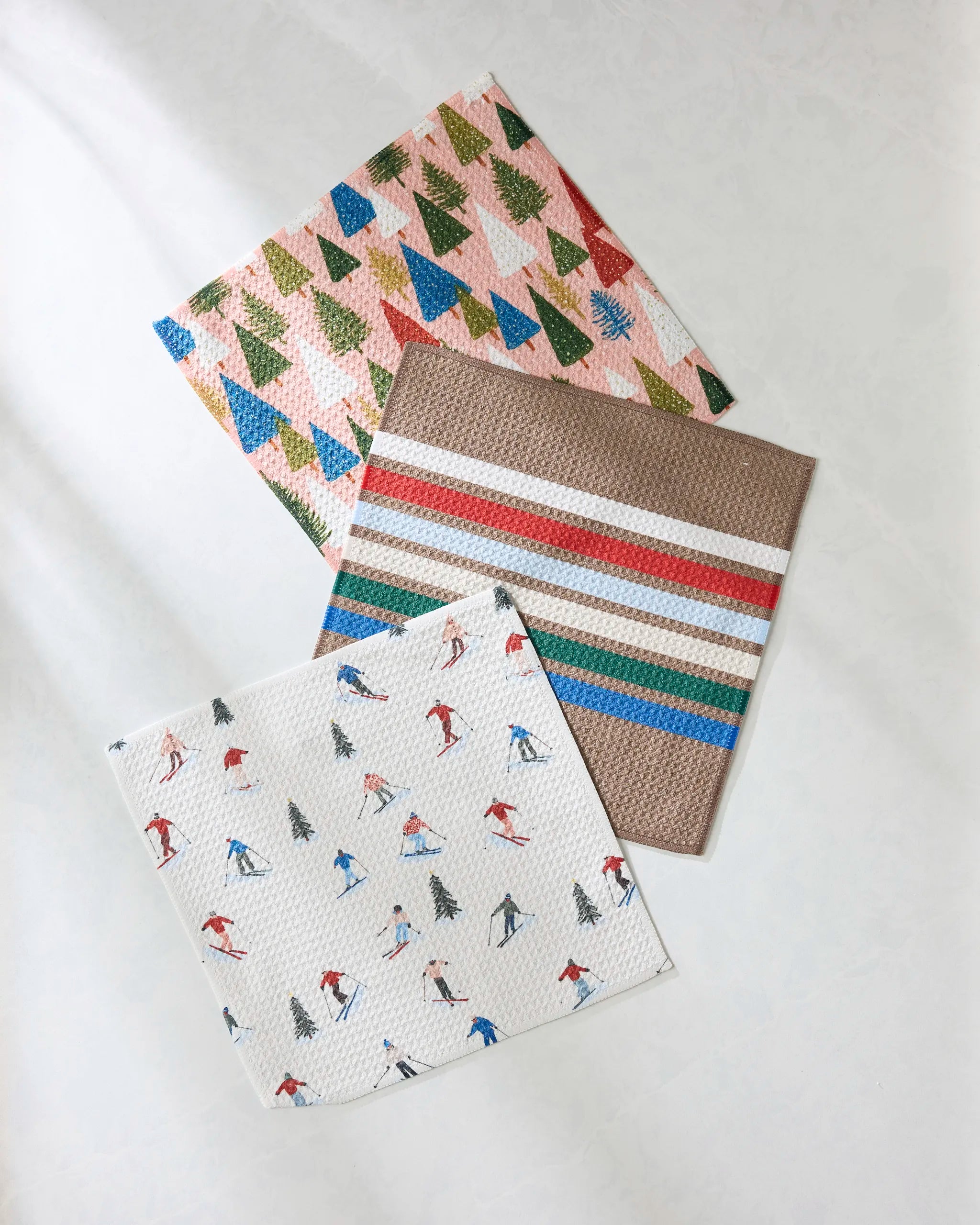 Three Geometry Winterhaus dishcloths are displayed on a light surface: one with a colorful tree pattern, one with multi-colored stripes, and one with skiers and trees on a white background.