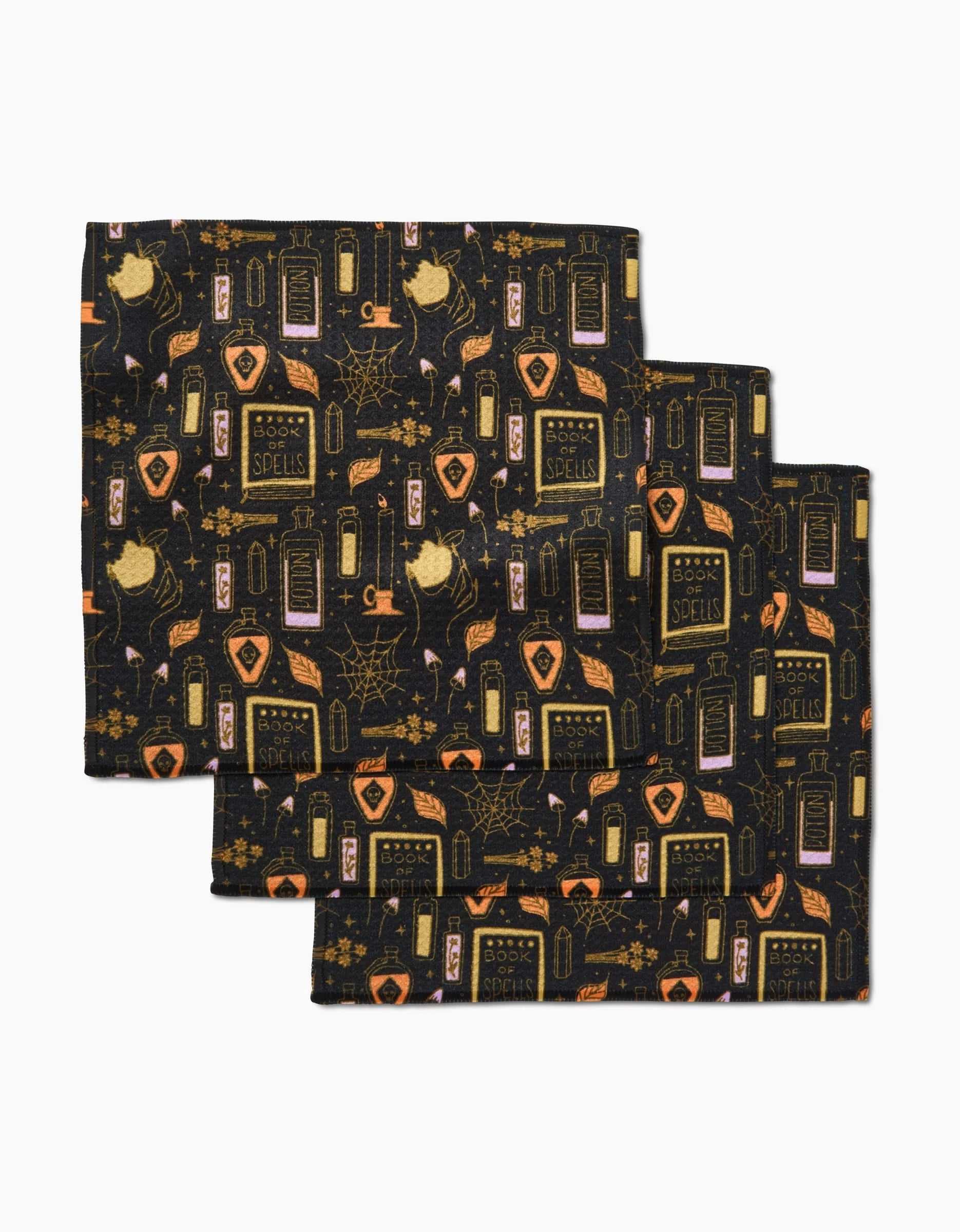 Three folded black cloth napkins from the Mandy Arroyo Witchy Things set feature a Halloween pattern with spell books, cats, potions, cobwebs, candles, and mystical items in orange, yellow, and pink tones. Super-absorbent dishcloth set.