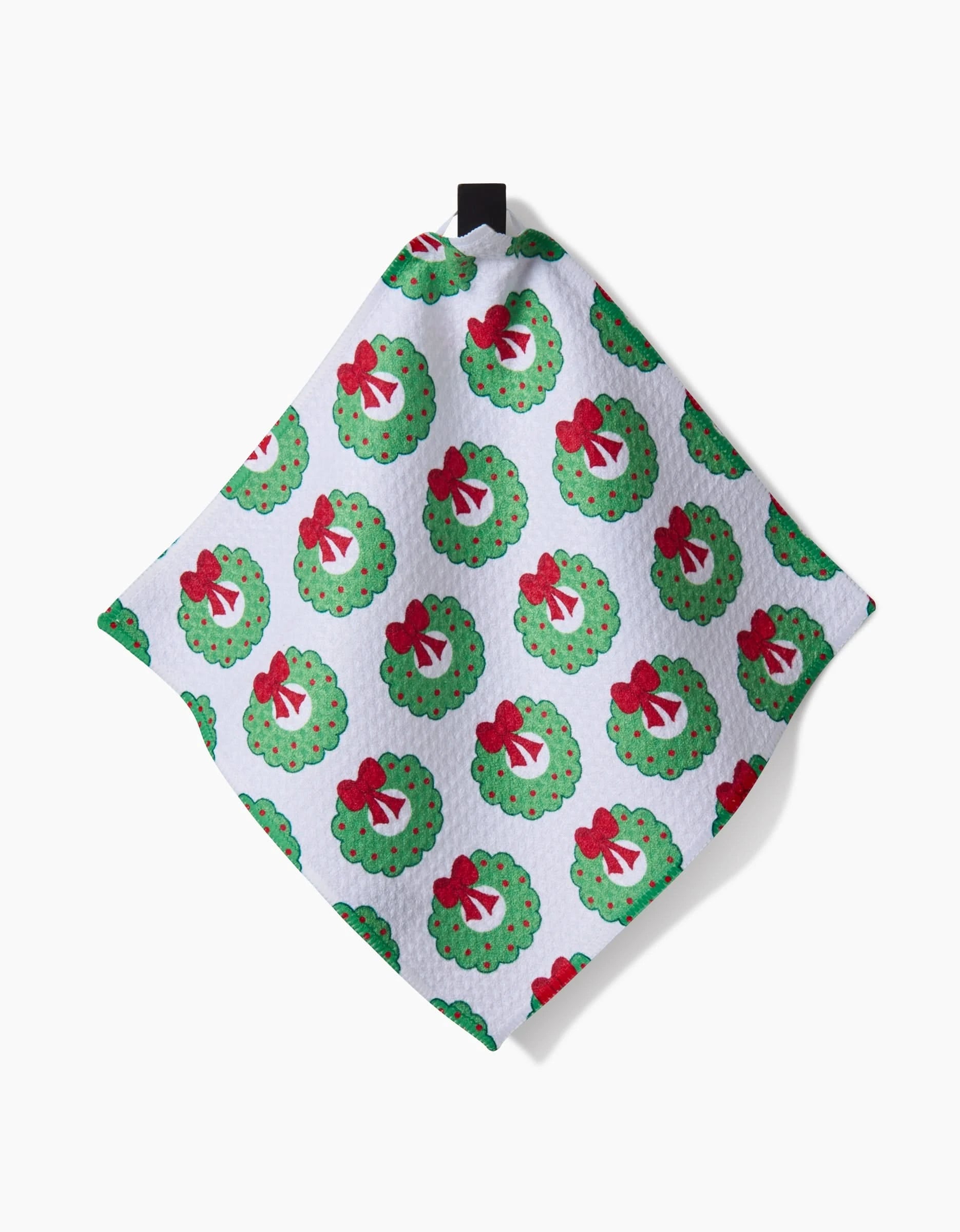 Mindfulnice’s Wreath Wishes is a white textured dishcloth set with green Christmas wreaths, red bows, and Santa faces in a repeating pattern, and a black hanging loop at the top.