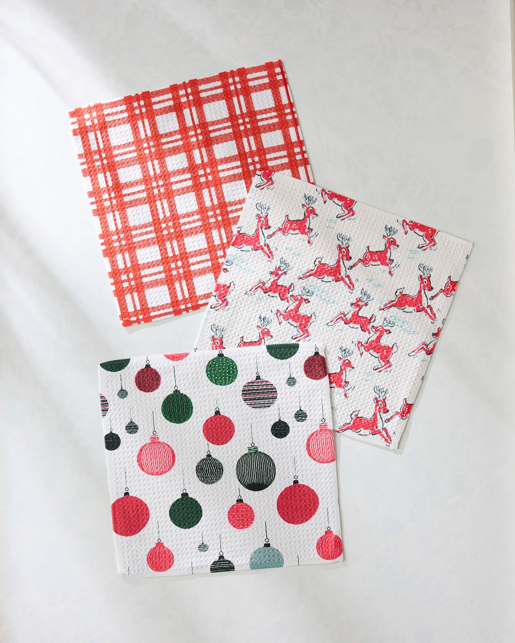 Geometry’s Xmas Day set features three recycled square dishcloths on a white surface: one red plaid, one with red reindeer, and one adorned with red, green, and black Christmas ornaments.