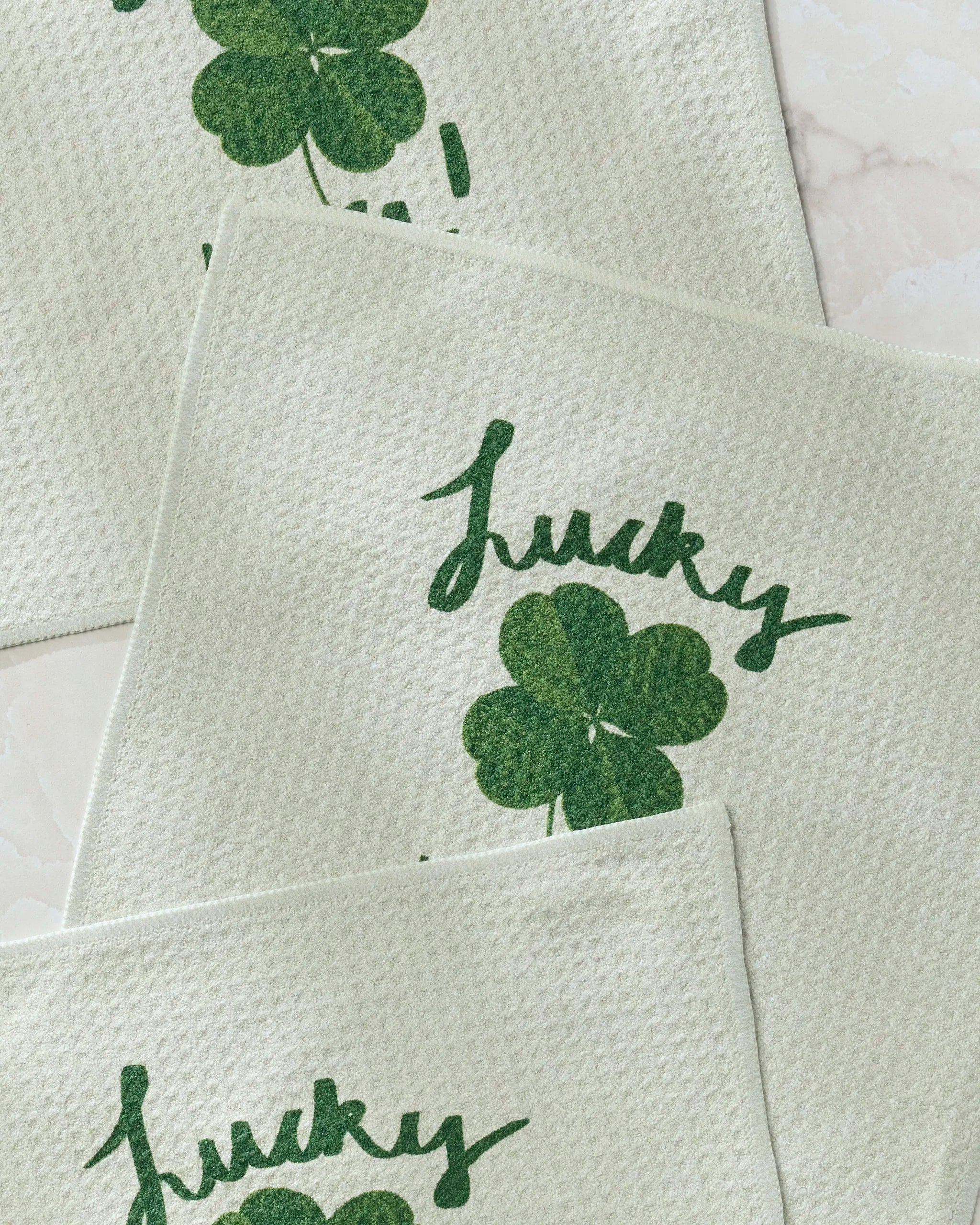 Three light-colored Geometry "Your Day" dishcloths, each displaying a large green four-leaf clover and the word "Lucky" in green cursive, are arranged on a light marble surface.