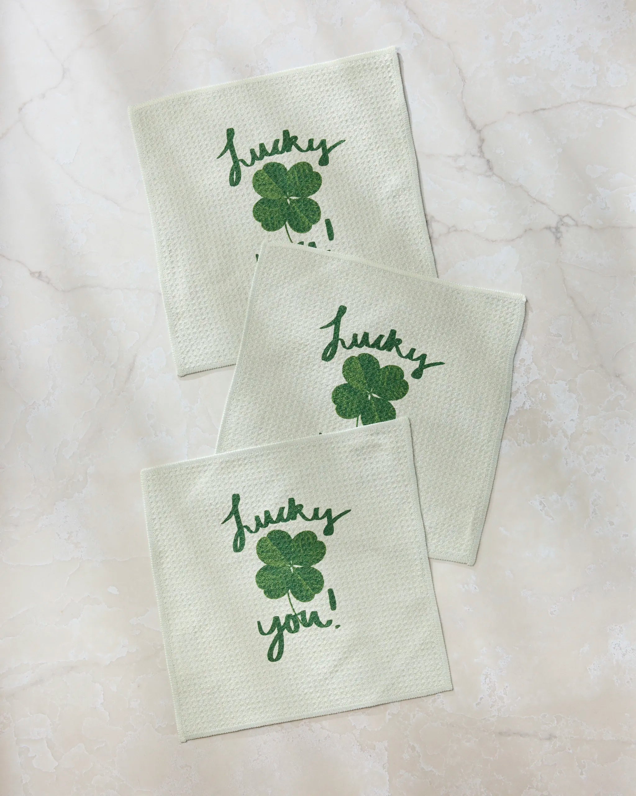 Three "Your Day" napkins by Geometry, made from recycled materials, feature green four-leaf clovers and the phrase "Lucky you!" in green script. Arranged in a loose triangle on a marble surface.