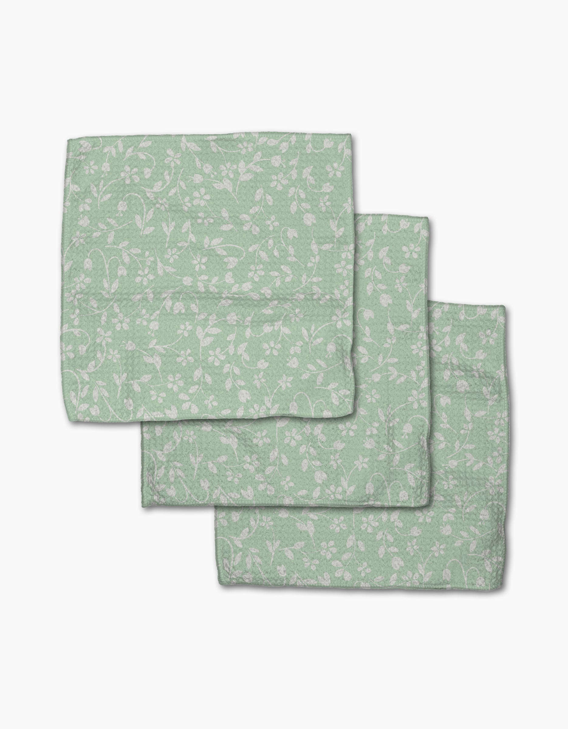 Three folded Dandy napkins by GEOMETRY, in light green recycled fabric with a subtle white floral and vine pattern, are arranged overlapping on a white background.