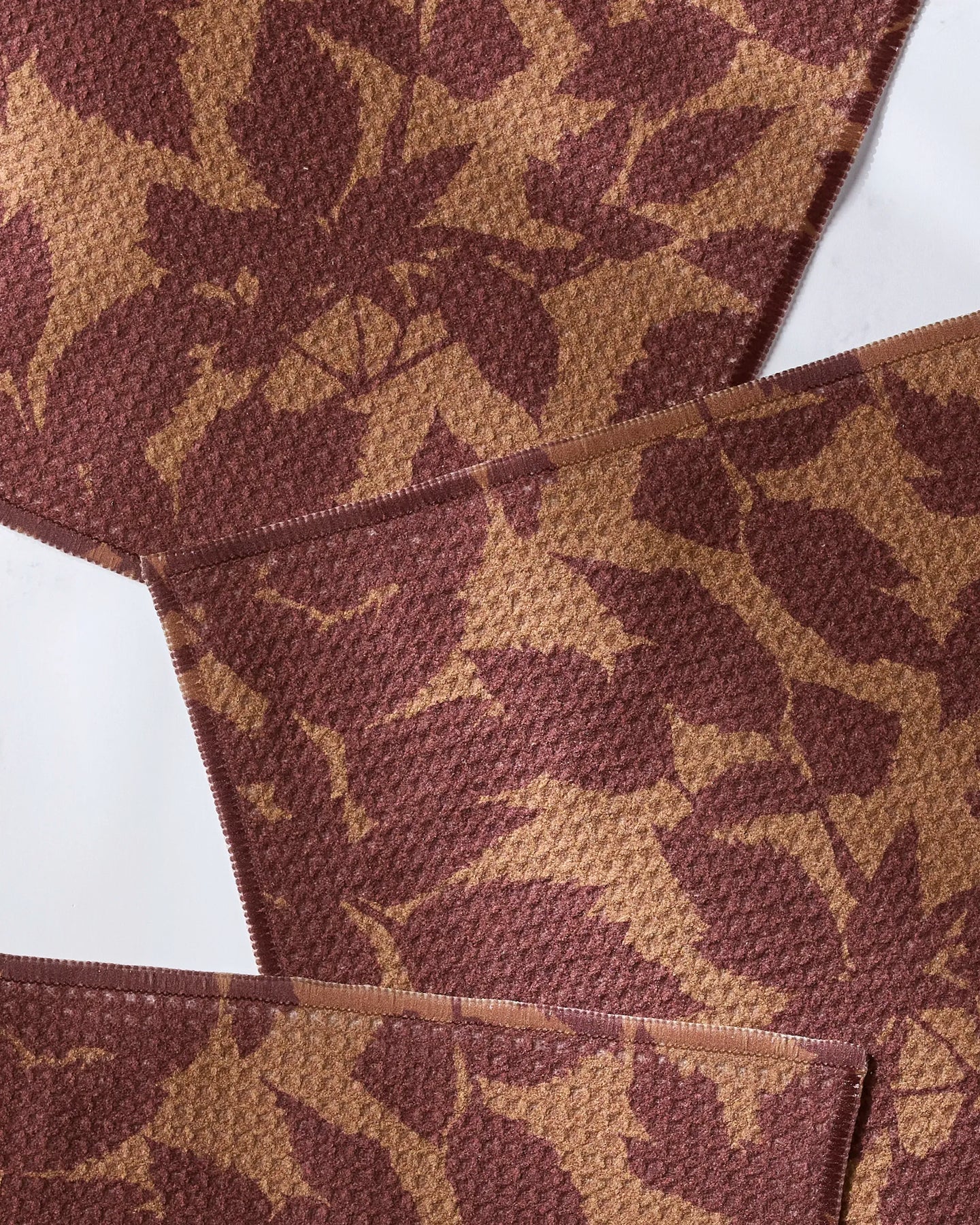 Close-up of Rustle by Geometry, featuring overlapping fabric swatches with a textured surface and brown and tan leaf patterns, crafted from recycled materials and arranged diagonally on a white background.