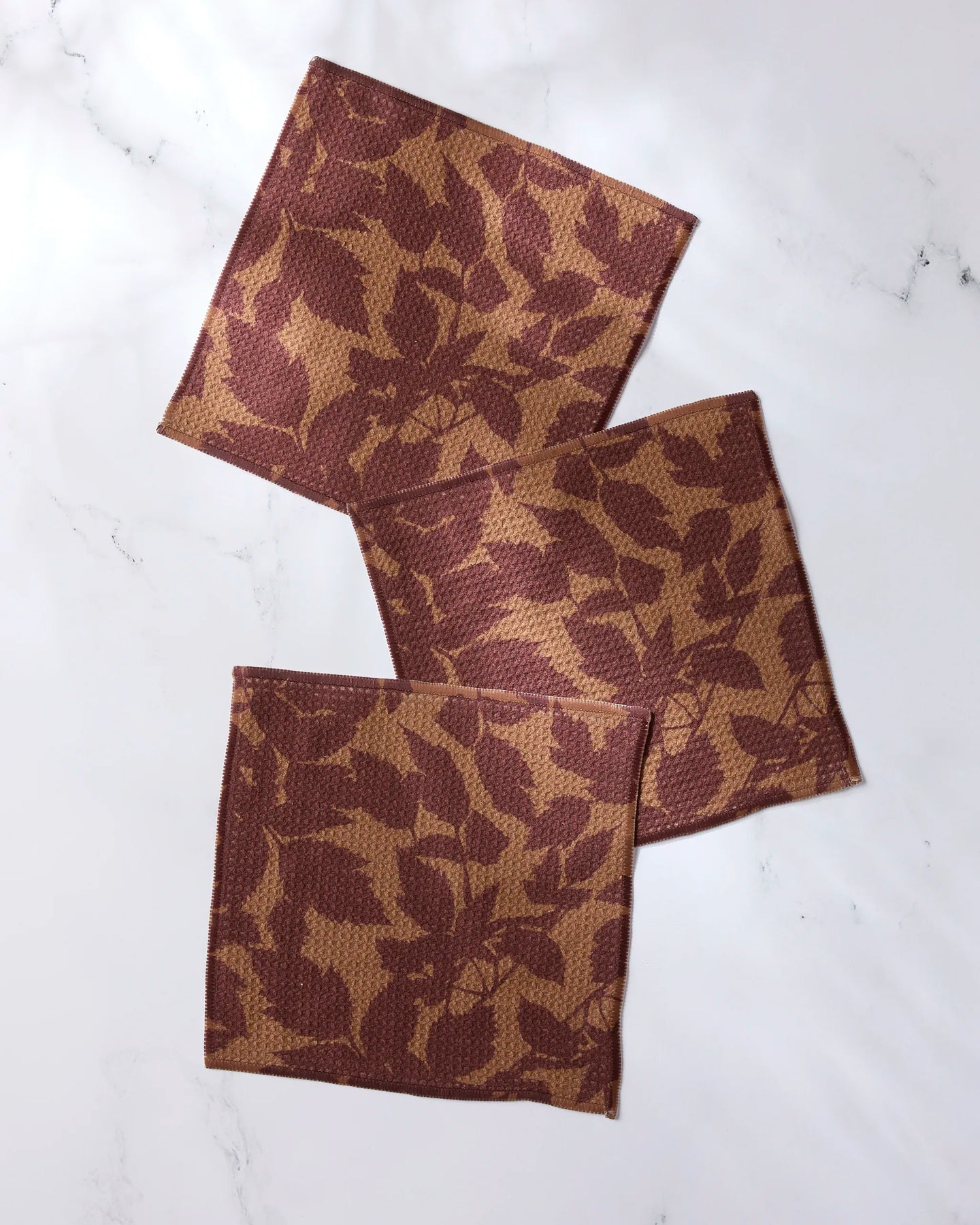 Three Rustle square cloth napkins by Geometry, featuring a brown and tan leaf pattern and made from recycled materials, are arranged overlapping on a white marble surface.