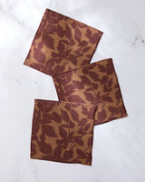 Three Rustle square cloth napkins by Geometry, featuring a brown and tan leaf pattern and made from recycled materials, are arranged overlapping on a white marble surface.