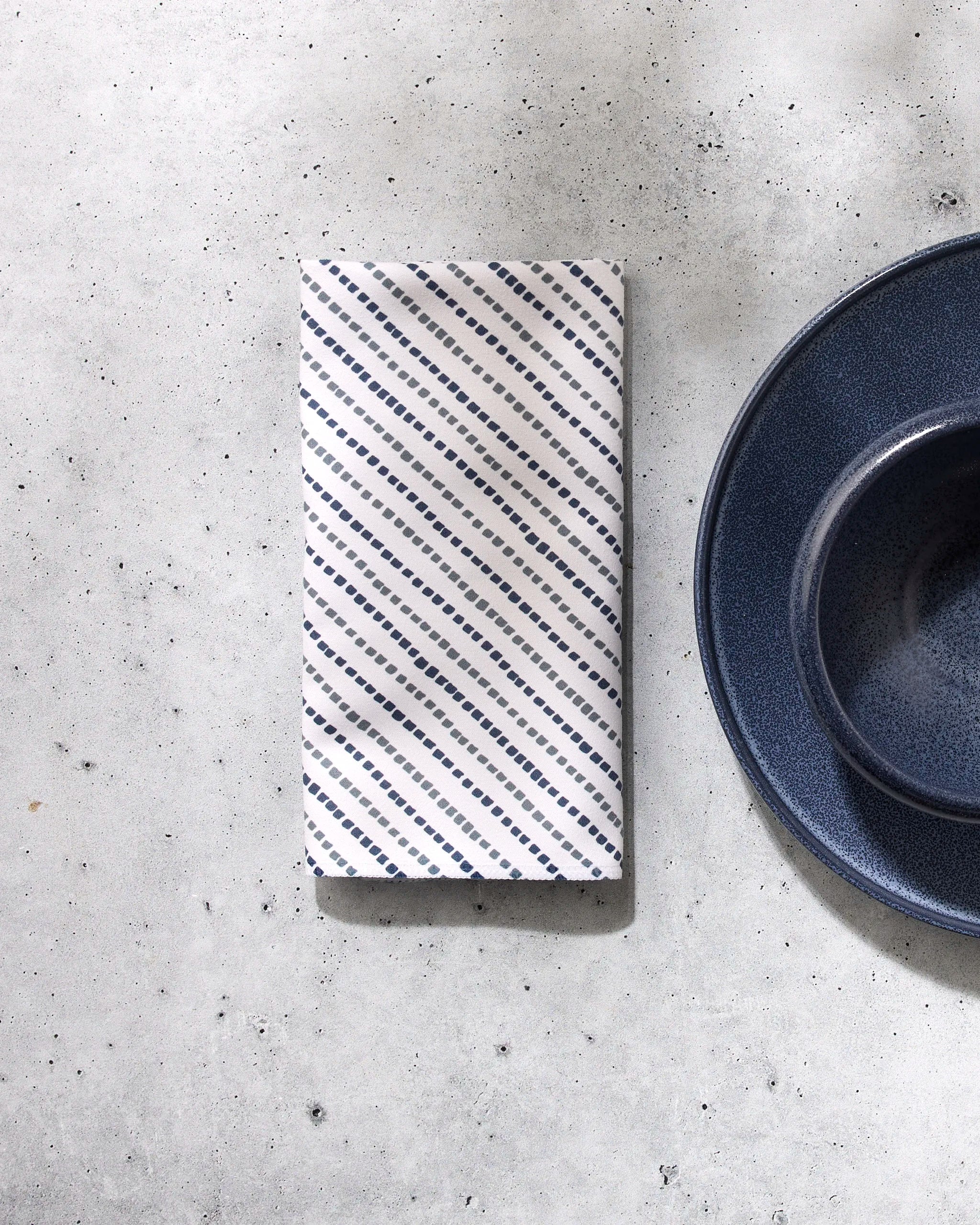 A folded, patterned white Kealia reusable table napkin by GEOMETRY rests on a light gray speckled surface beside a partially visible dark blue ceramic plate and bowl to the right.