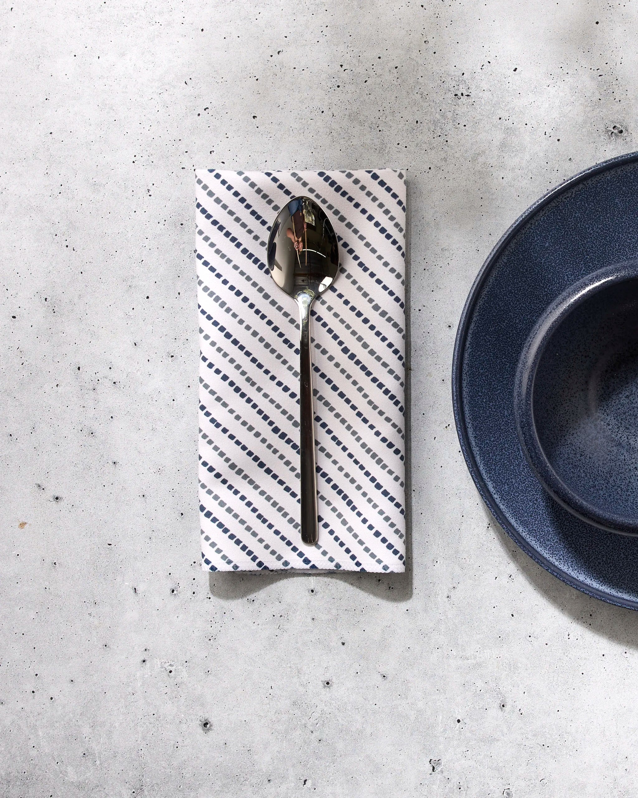 A GEOMETRY Kealia silver spoon rests on a white reusable napkin with blue diagonal stripes, set on a light gray surface beside a dark blue plate and bowl.