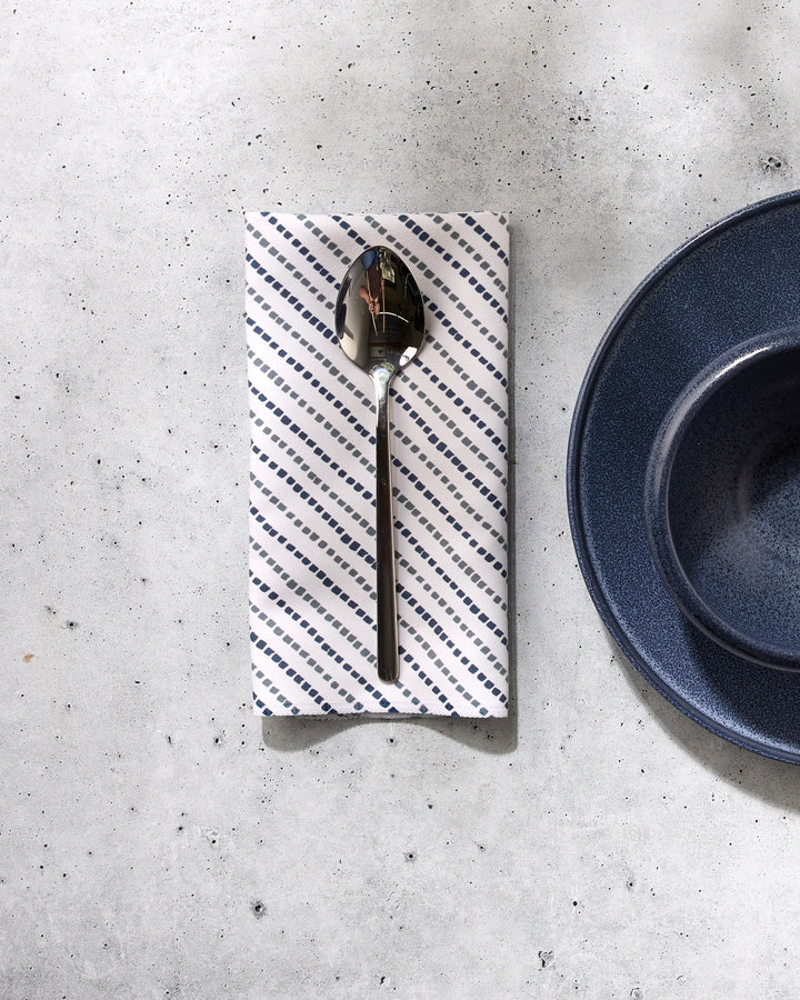 A GEOMETRY Kealia silver spoon rests on a white reusable napkin with blue diagonal stripes, set on a light gray surface beside a dark blue plate and bowl.