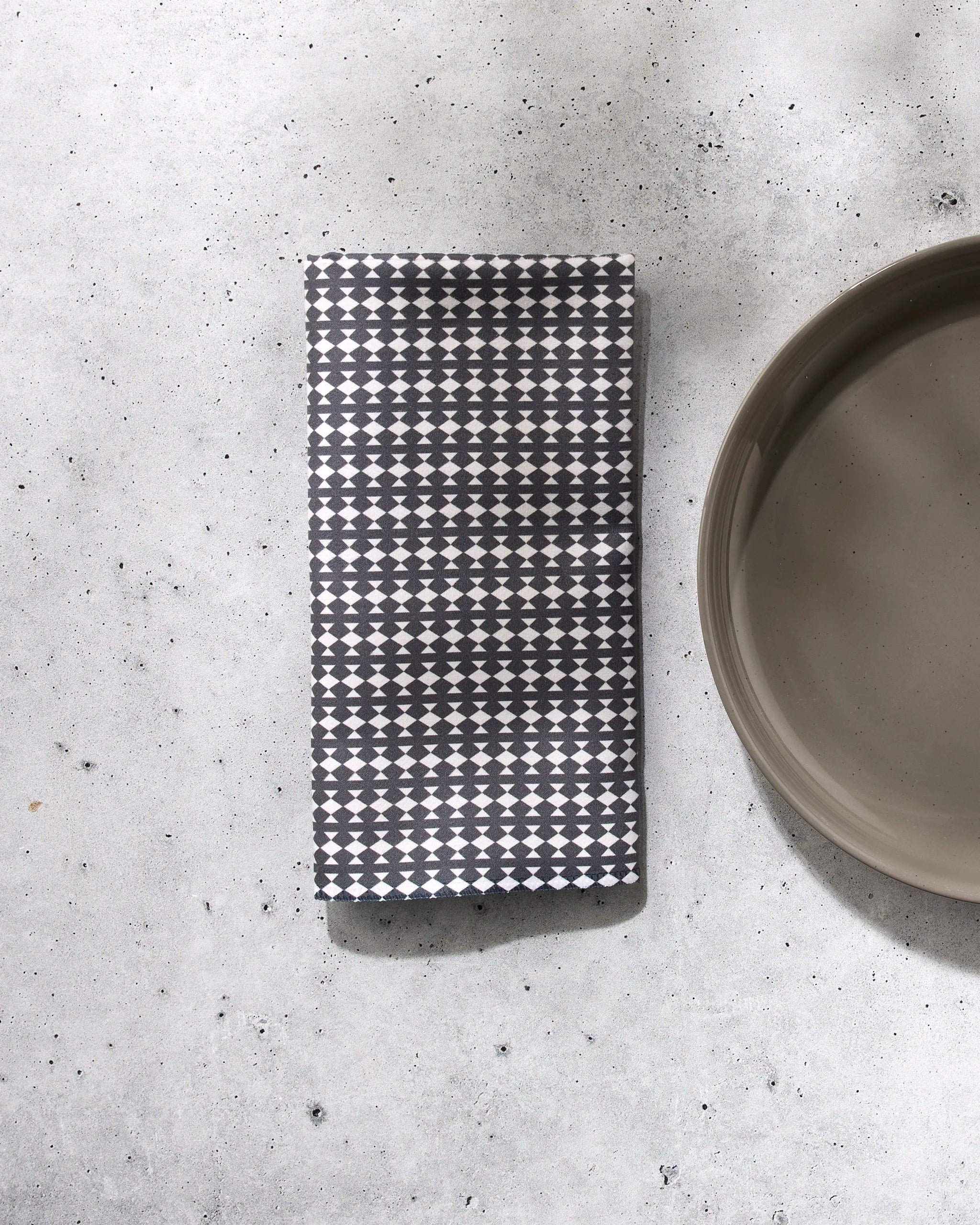 A folded reusable Maori napkin by GEOMETRY, featuring a black and white geometric pattern, rests on a light gray speckled surface beside a round beige plate.