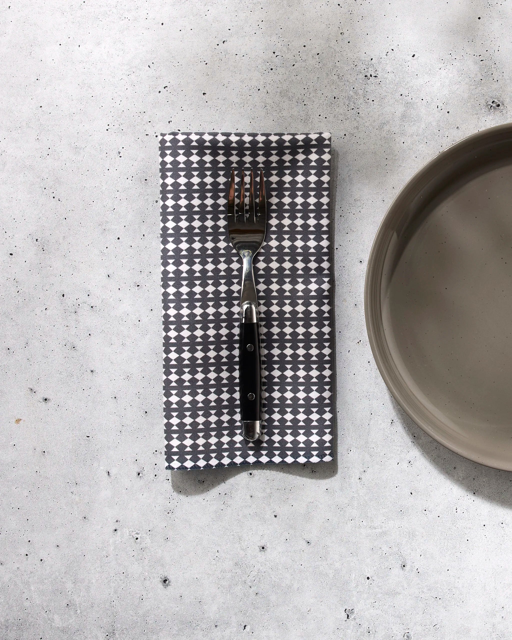 A fork rests on a folded Maori patterned gray dinner napkin by GEOMETRY, set next to a round light gray plate on a speckled light surface.