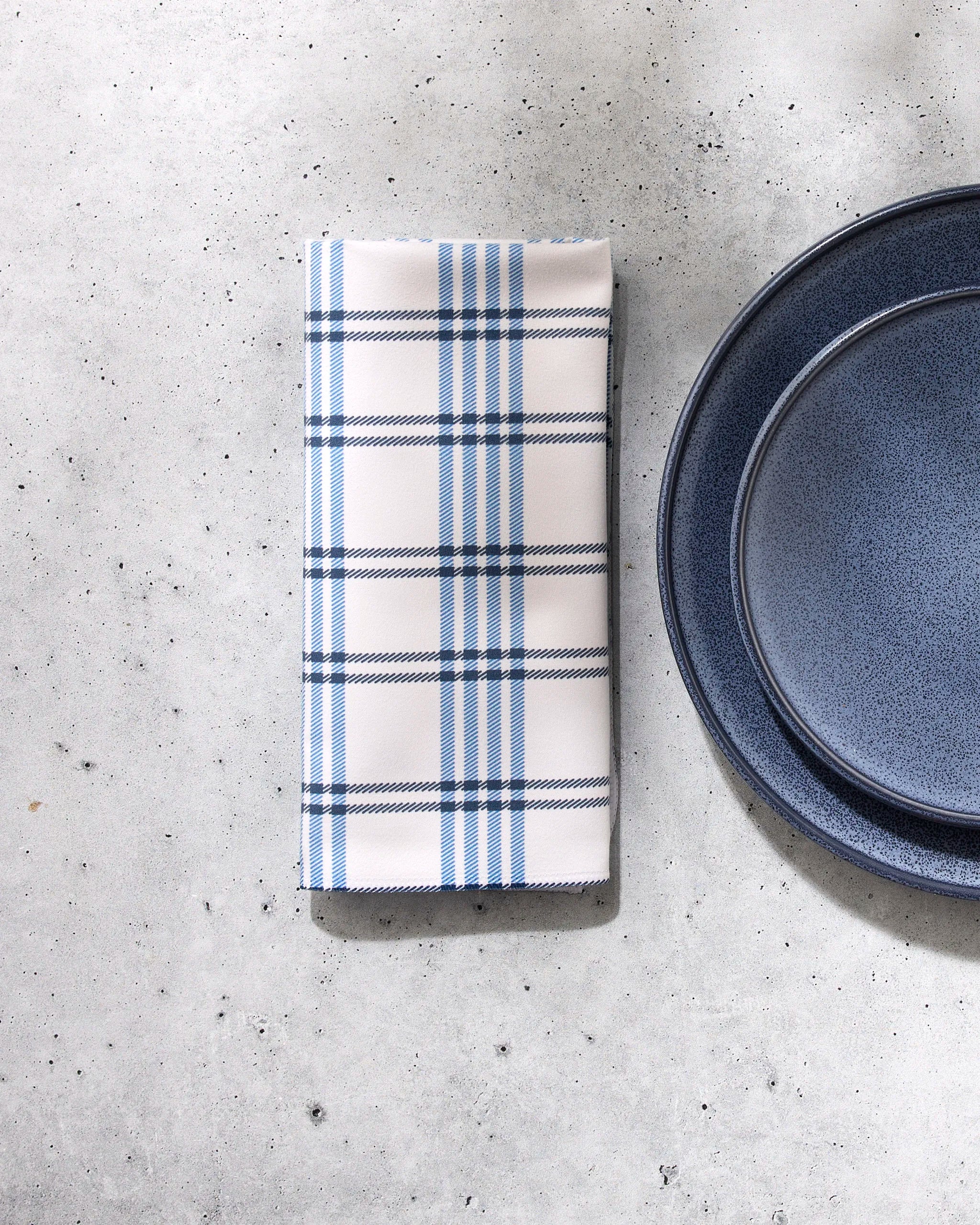 The GEOMETRY Seafaring napkin, folded and white with blue plaid stripes, rests beside two stacked blue plates on a light gray speckled surface.