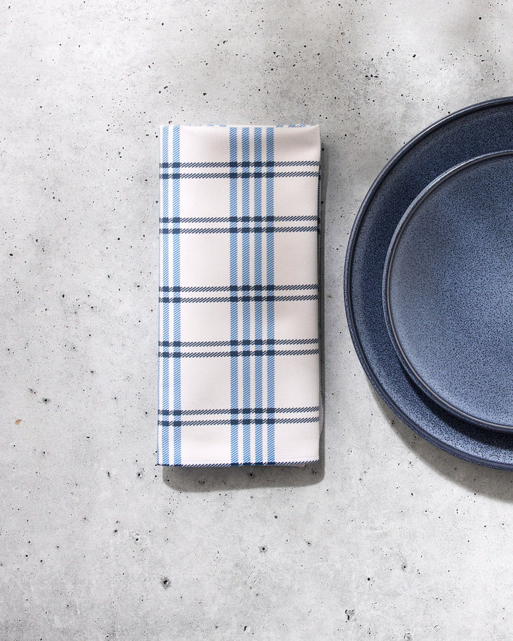 The GEOMETRY Seafaring napkin, folded and white with blue plaid stripes, rests beside two stacked blue plates on a light gray speckled surface.