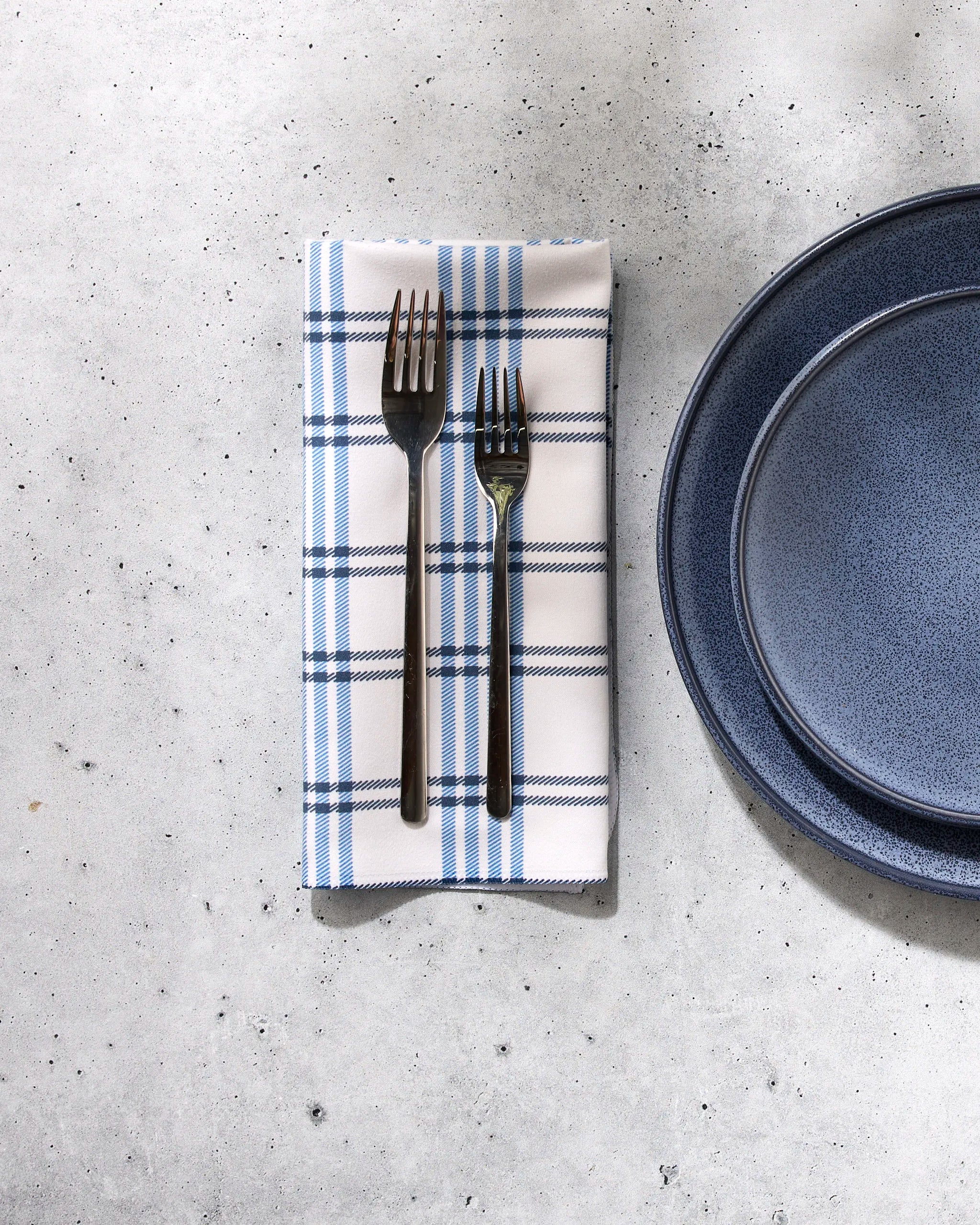 Two dark metal forks lie on a folded blue-and-white checked Seafaring napkin by GEOMETRY, placed next to stacked dark blue ceramic plates, all arranged on a light gray stone surface.
