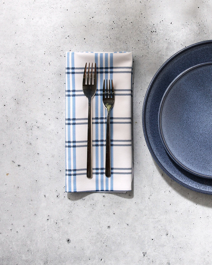 Two dark metal forks lie on a folded blue-and-white checked Seafaring napkin by GEOMETRY, placed next to stacked dark blue ceramic plates, all arranged on a light gray stone surface.