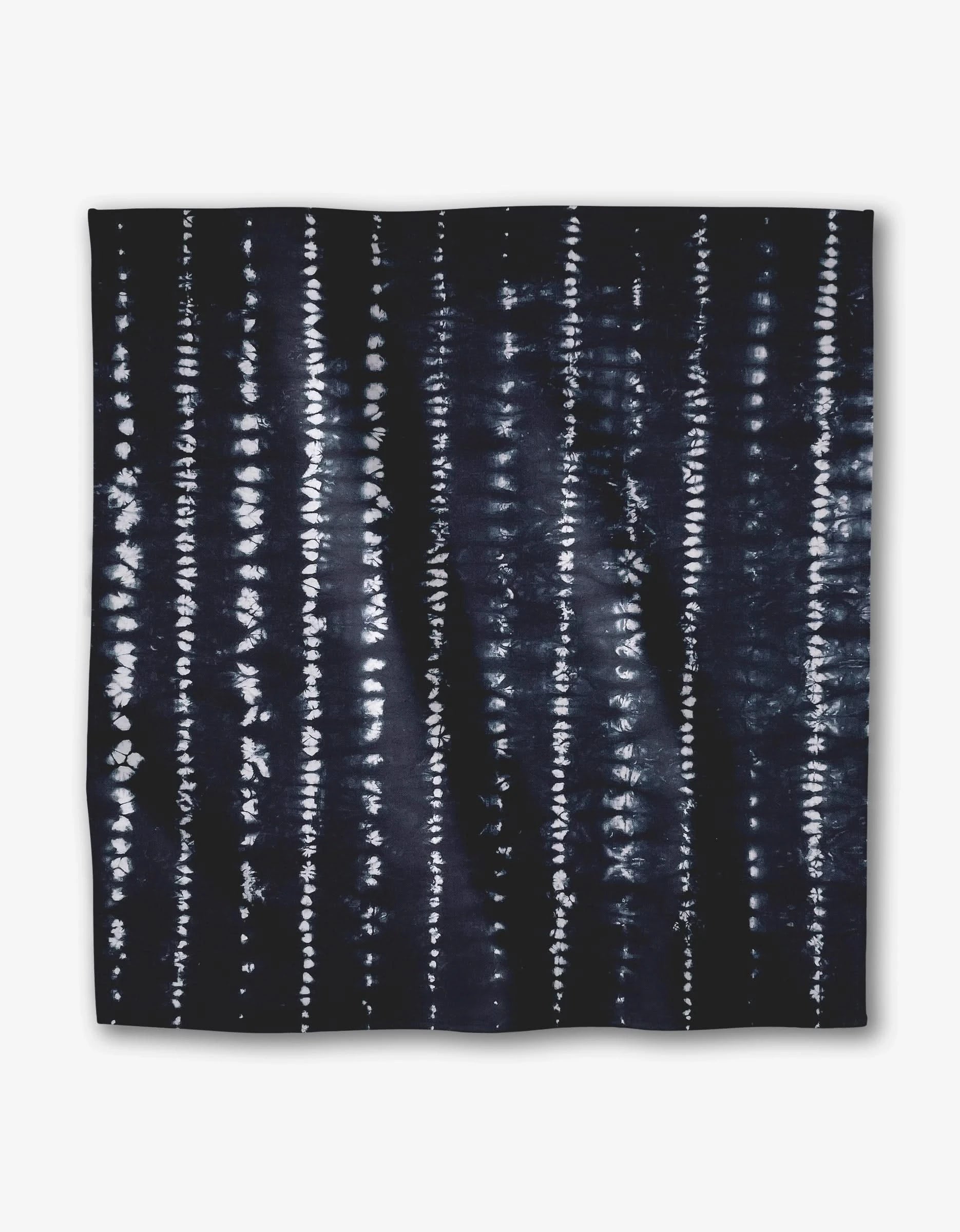 The Shibo dinner napkin set by GEOMETRY features a square design in Geosuede fabric with a dark indigo base, accented by vertical white tie-dye stripes. Made from recycled materials for a textured, eco-friendly look.