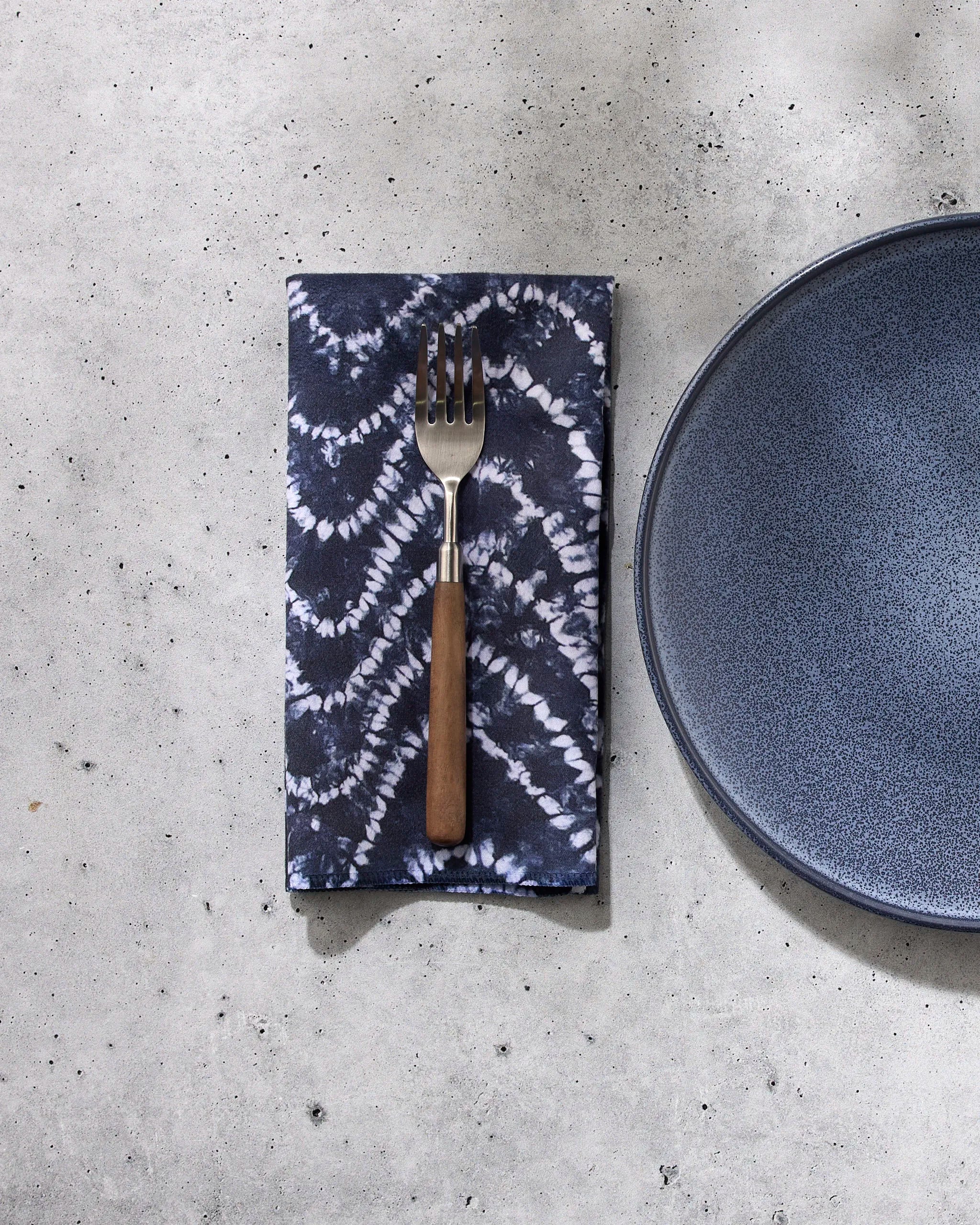 A GEOMETRY Shibo fork with a wooden handle rests on a folded, dark blue patterned reusable napkin beside a blue ceramic plate, all set on a light gray speckled surface.