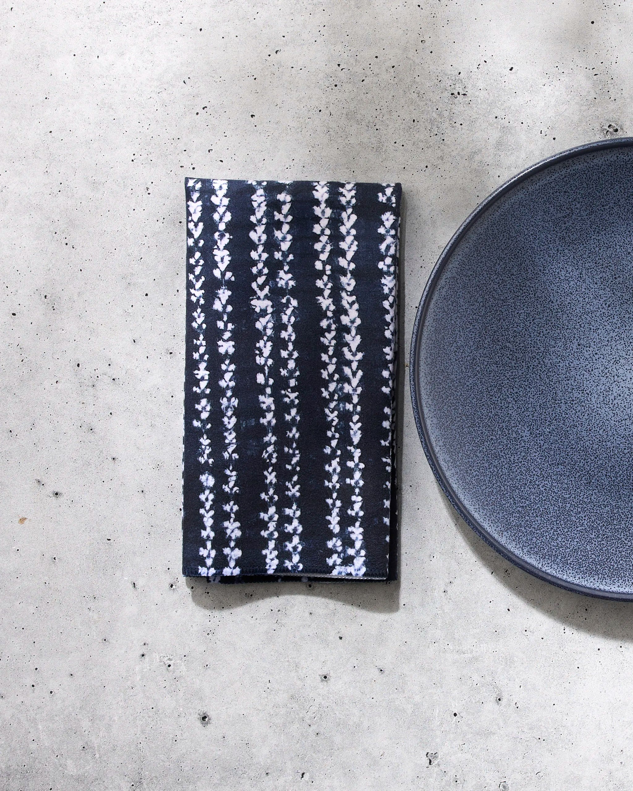The GEOMETRY Shibo reusable table napkin, featuring a white floral pattern, is neatly folded beside a dark gray ceramic plate on a light gray textured surface.