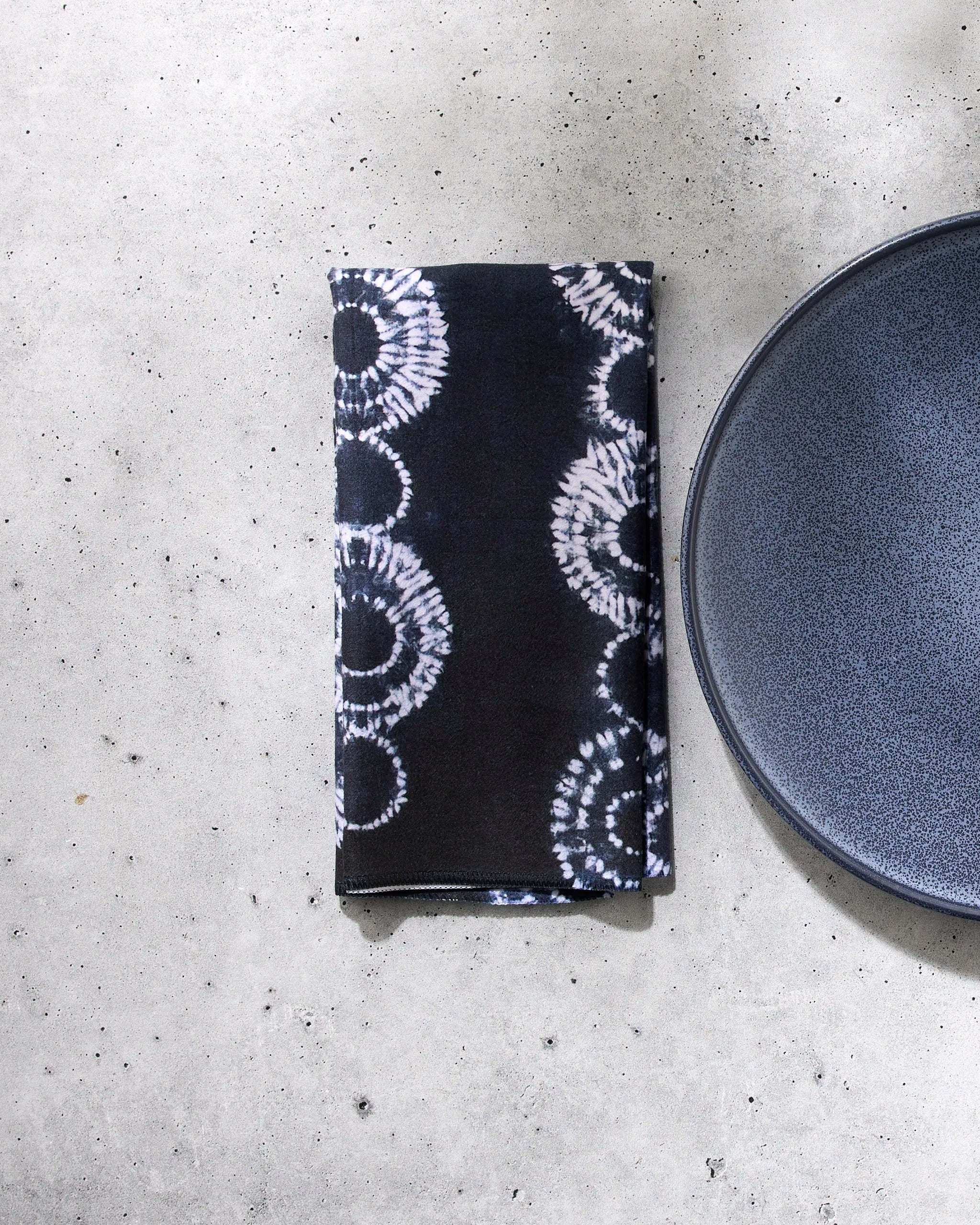 The Shibo napkin by GEOMETRY, featuring a black reusable fabric with a white circular tie-dye pattern, is displayed next to a dark blue plate on a light gray concrete surface.