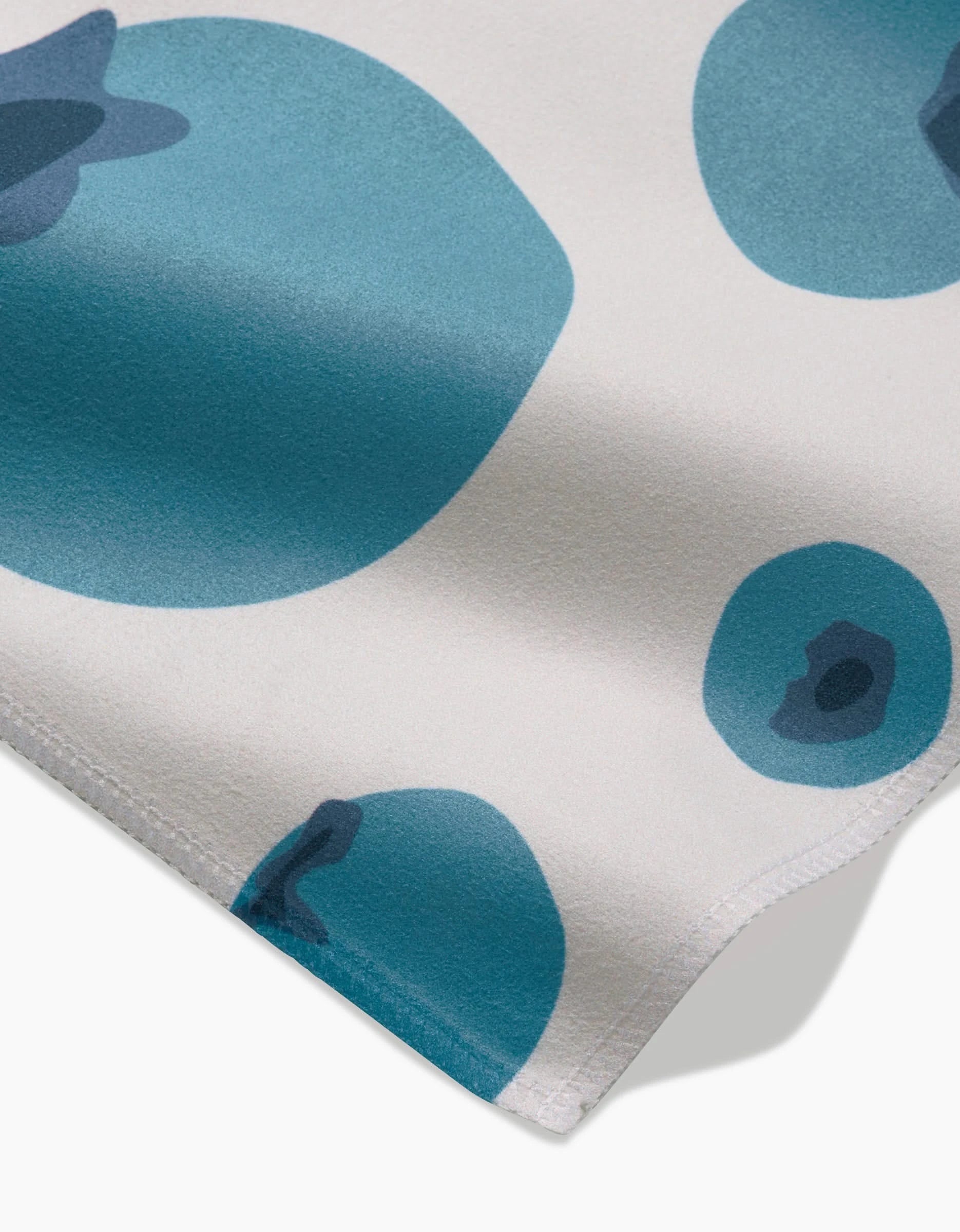 A close-up of GEOMETRYs Sunny Fruit shows white Geosuede fabric with large and small abstract blue circles—some with dark details—draped elegantly in the corner, ideal for a stylish Dinner Napkin Set.
