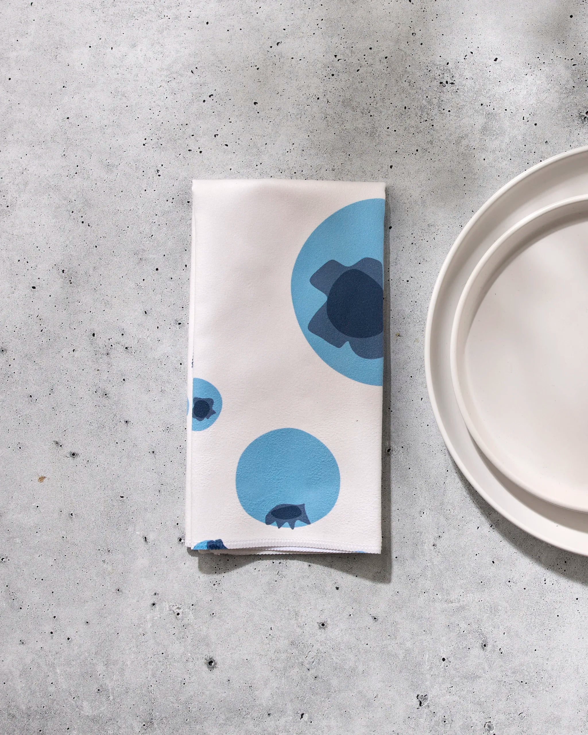 A folded white GEOMETRY napkin, "Sunny Fruit," featuring blue circular patterns, rests on a textured gray surface beside a white plate.