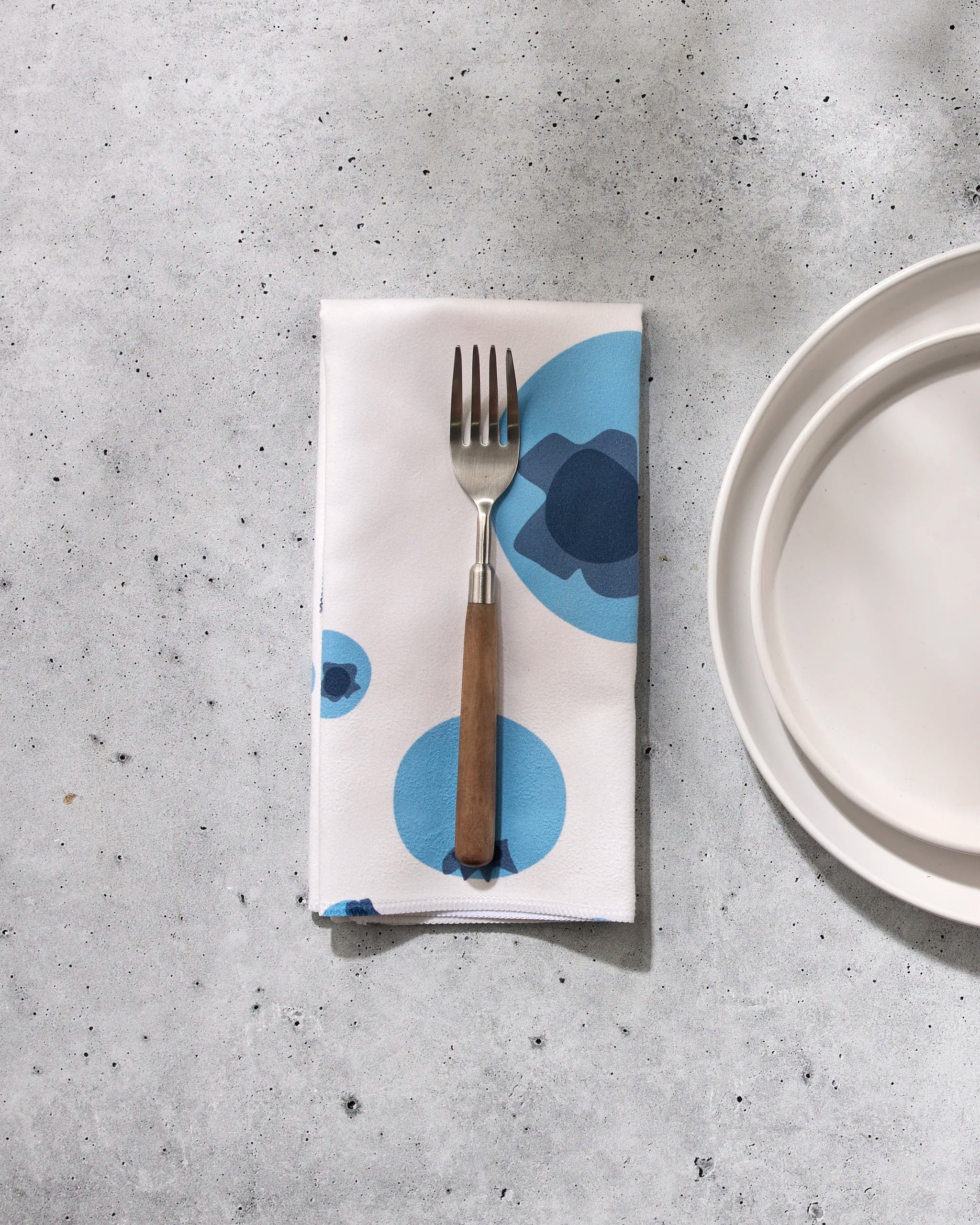 A GEOMETRY Sunny Fruit fork with a wooden handle sits on an eco-friendly napkin with blue circles, next to a white plate on a light gray speckled surface.