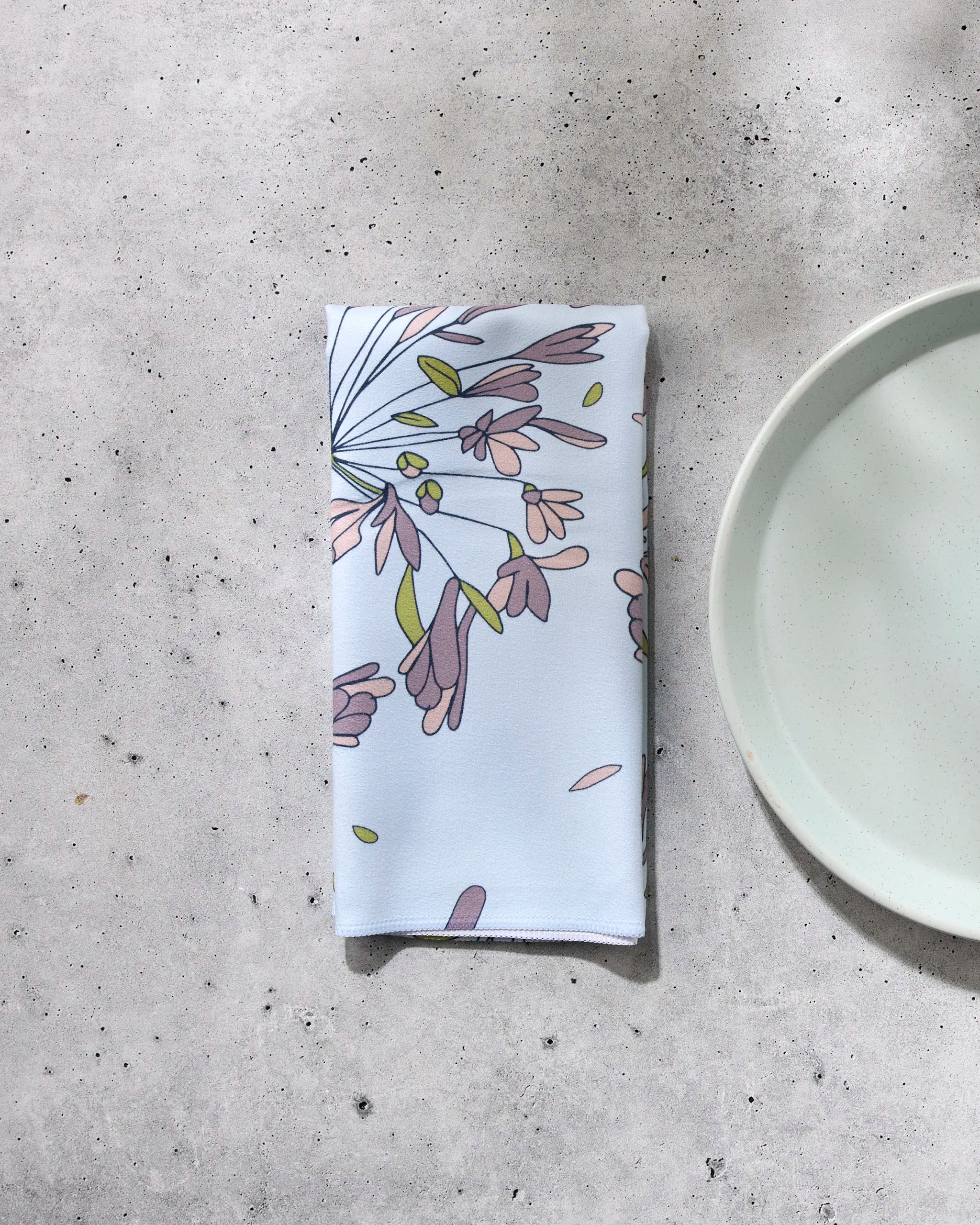 A folded GEOMETRY "The Wind" reusable napkin featuring a pastel floral pattern rests on a light gray textured surface beside a matching ceramic plate.