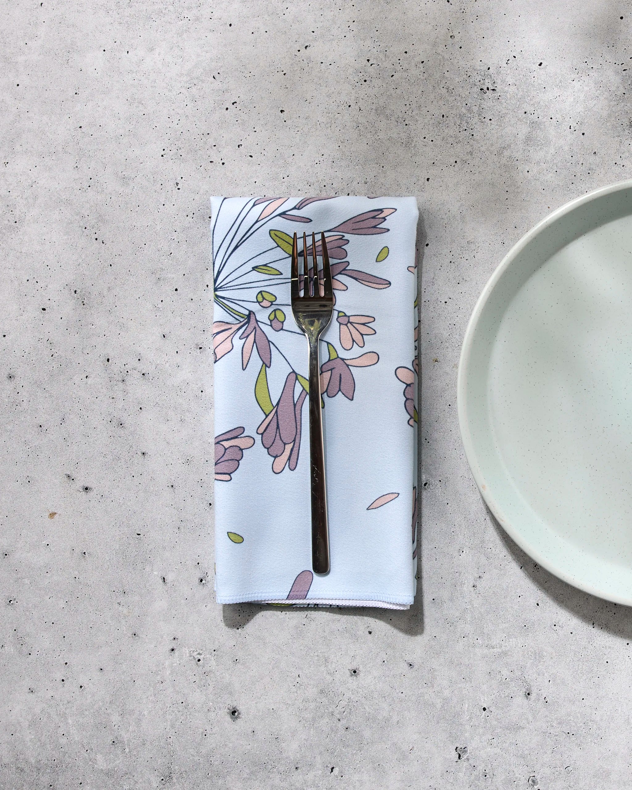 A silver fork rests on GEOMETRY's The Wind recycled floral-patterned dinner napkin set, next to an empty light green plate on a speckled gray surface.
