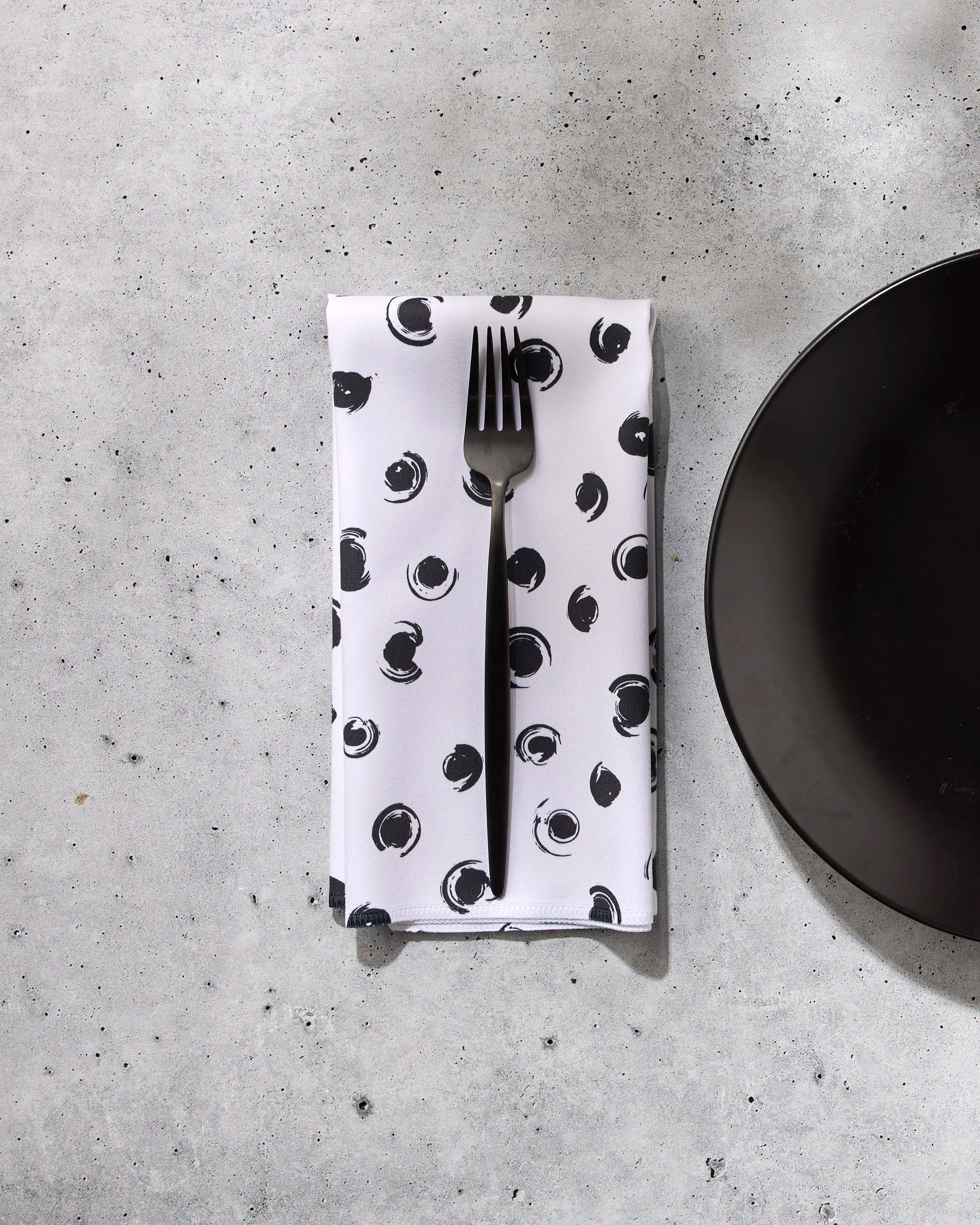 A black fork rests on a white GEOMETRY Tiny Drops recycled dinner napkin, featuring a black abstract dot pattern, placed on a light gray stone surface next to the edge of a plain black plate.
