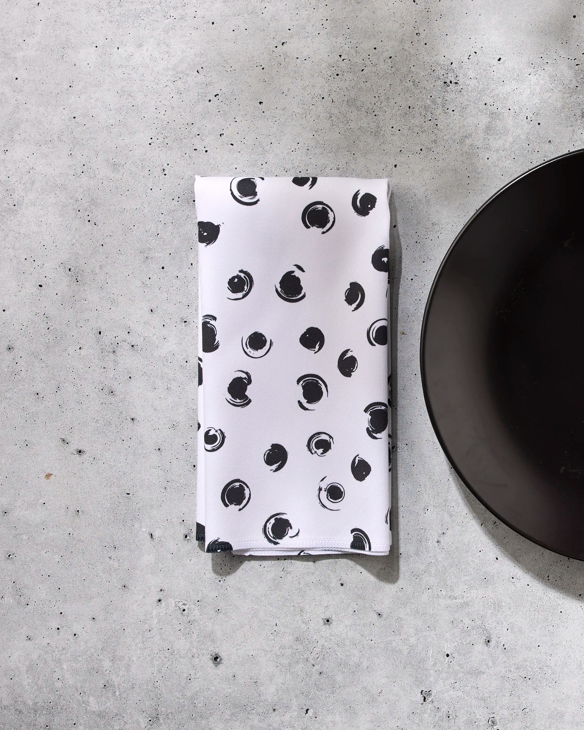 The GEOMETRY Tiny Drops napkin, featuring a black abstract dot pattern on white, is neatly folded on a light gray concrete surface beside the edge of a round black plate.