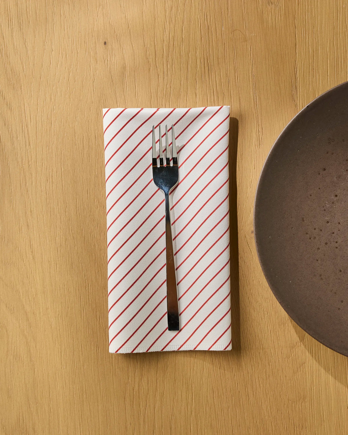 A fork rests on a Geometry Sugar Stripe reusable napkin with red diagonal stripes, placed on a light wooden table next to a brown plate.