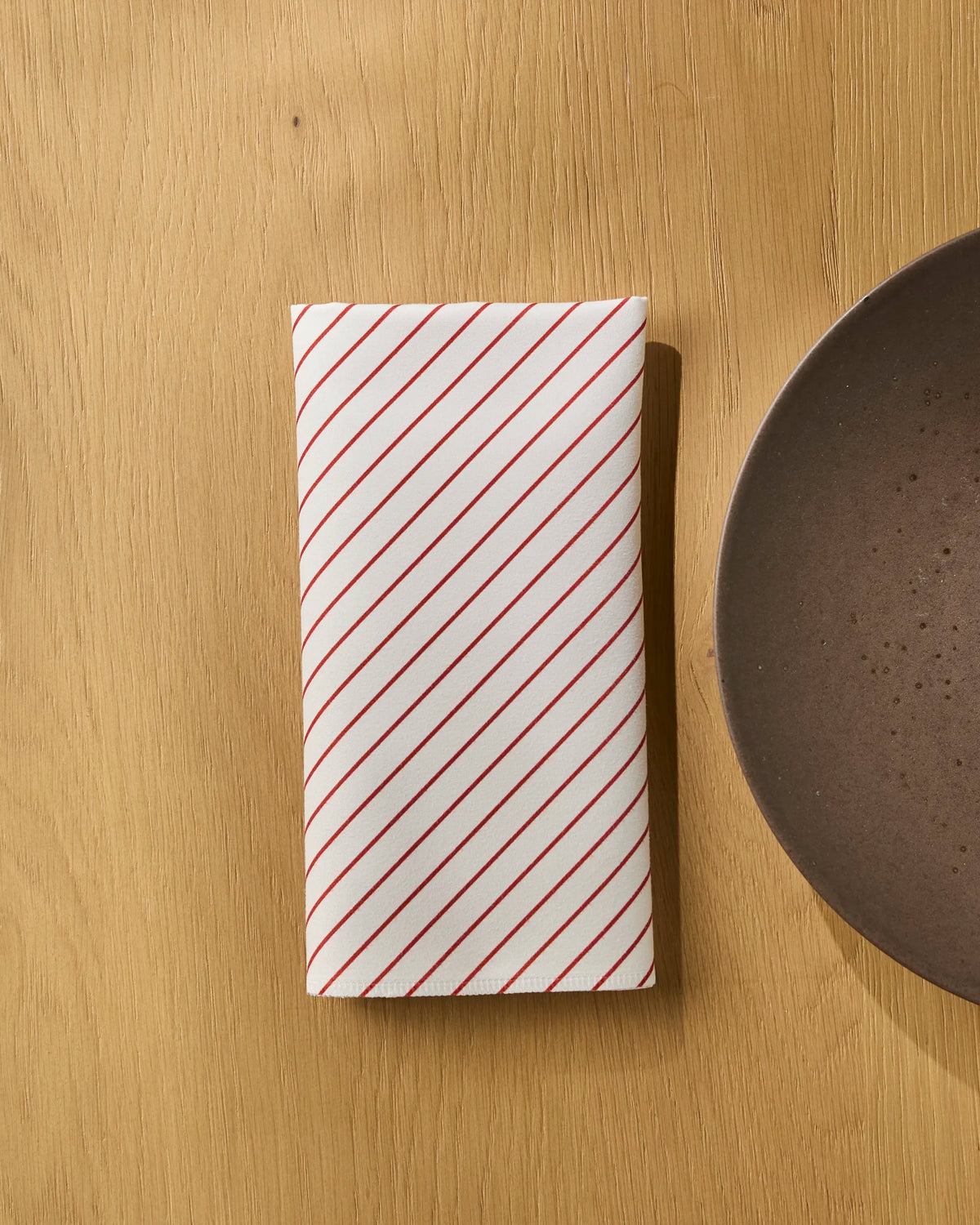 A Geometry Sugar Stripe folded white reusable napkin with diagonal red stripes rests on a light wooden surface next to a brown plate.