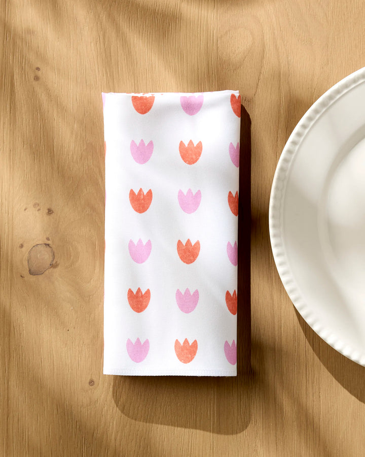 A folded white Addison dinner napkin by Amy MacCready, featuring pink and orange tulips, sits on a wooden table beside a scalloped plate. Sunlight and subtle shadows highlight these absorbent napkins.
