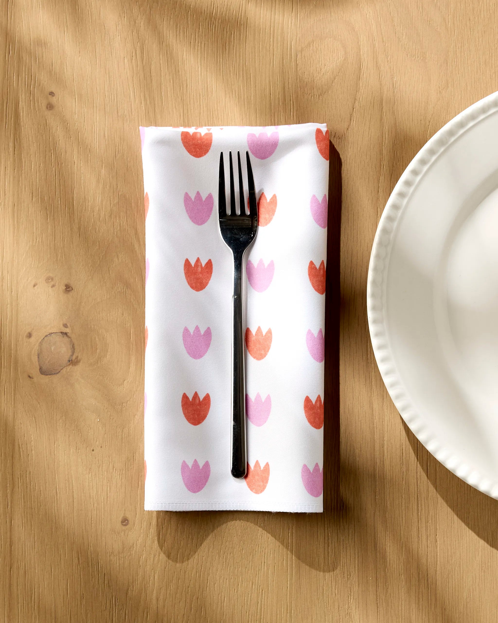 A metal fork rests on the Amy MacCready Addison Dinner Napkin Set with pink and red tulips, placed on a wooden table beside a white, scalloped-edge plate. Sunlight highlights these absorbent napkins’ vibrant design.