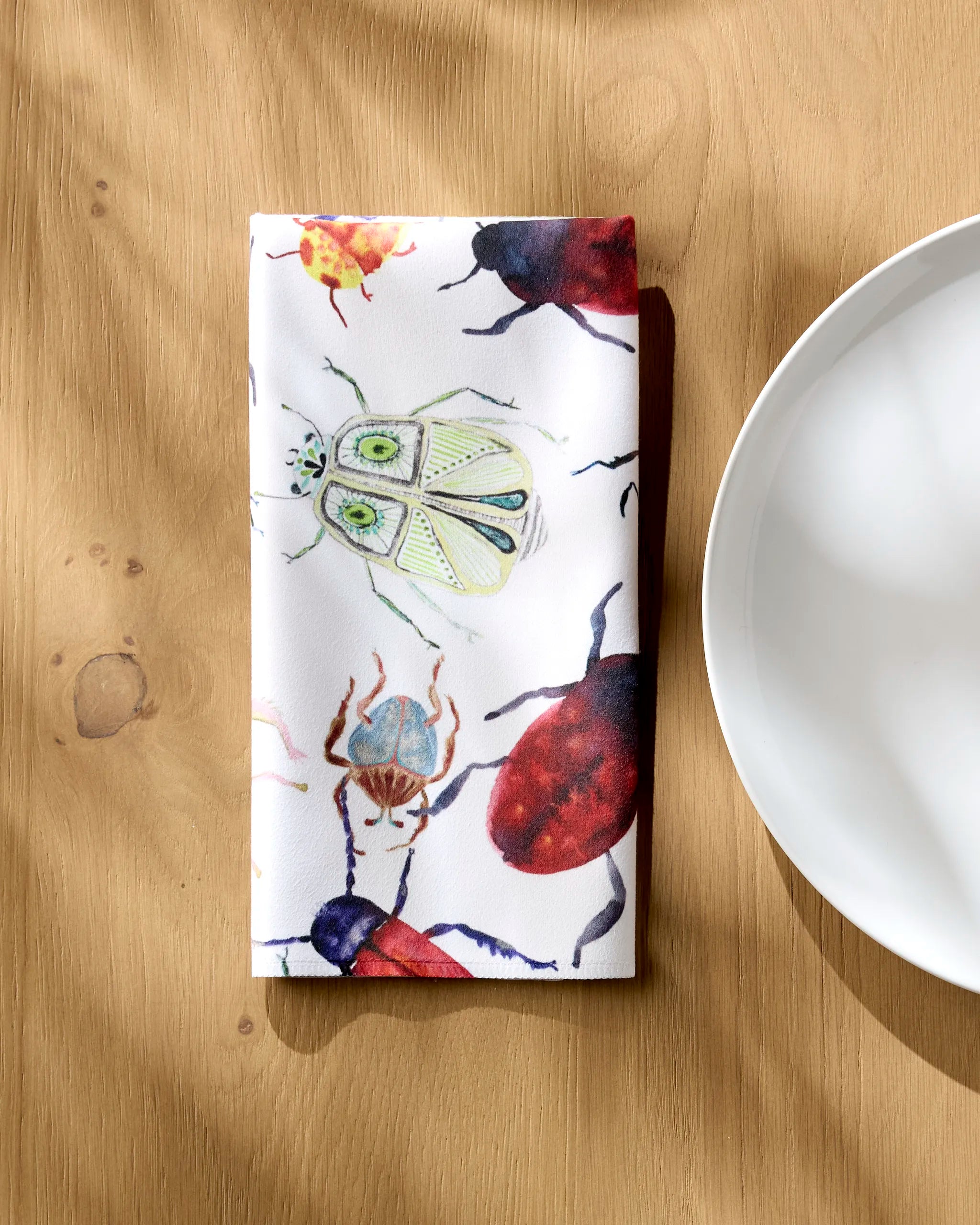A folded white reusable napkin featuring colorful African Beetles by Taryn Brennan rests on a wooden table beside a white plate, with sunlight casting gentle shadows across the scene.