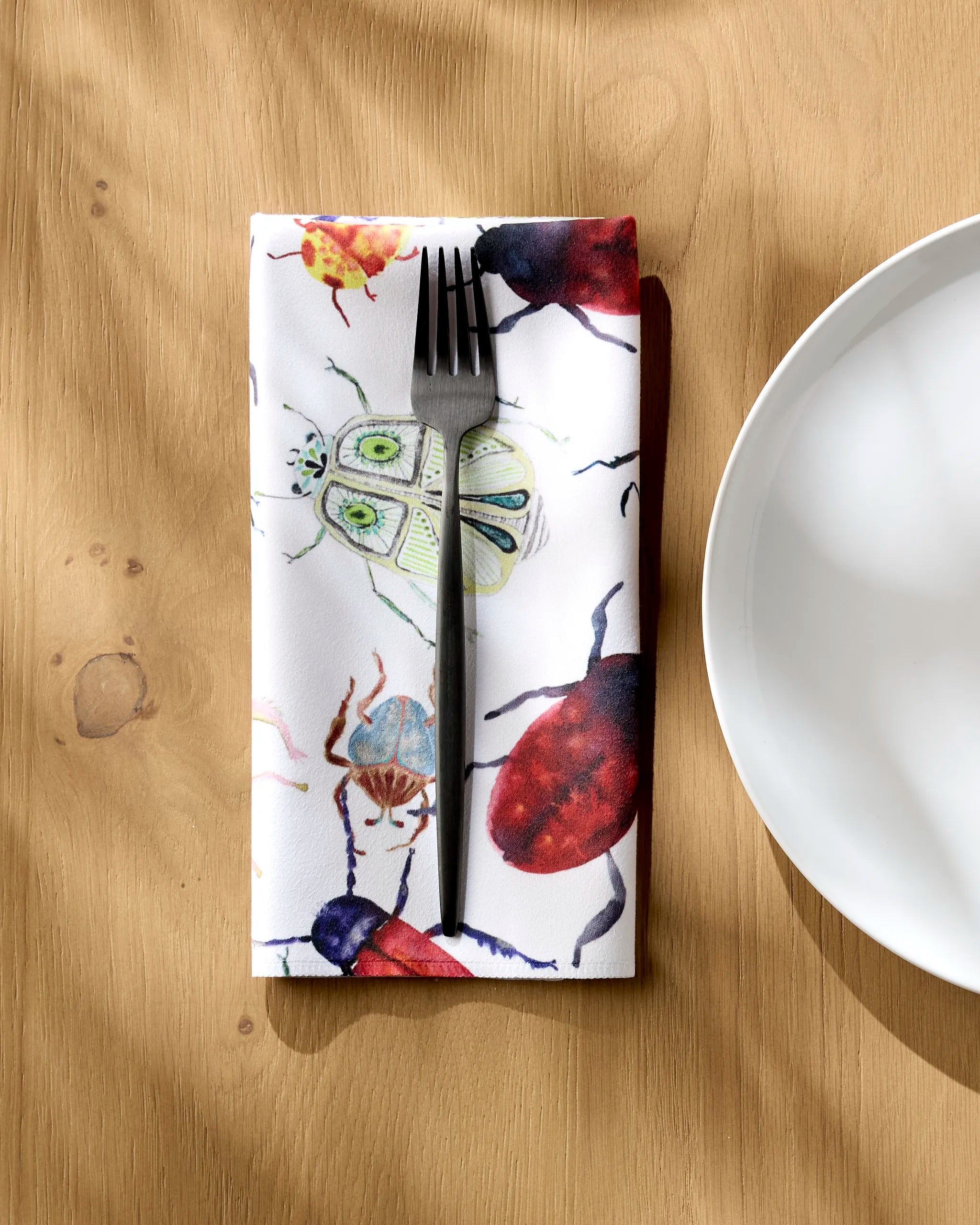 A fork rests on a Taryn Brennan "African Beetles" eco-friendly napkin featuring vibrant insect illustrations, placed beside a white plate on a light wood table as sunlight casts gentle shadows over the scene.