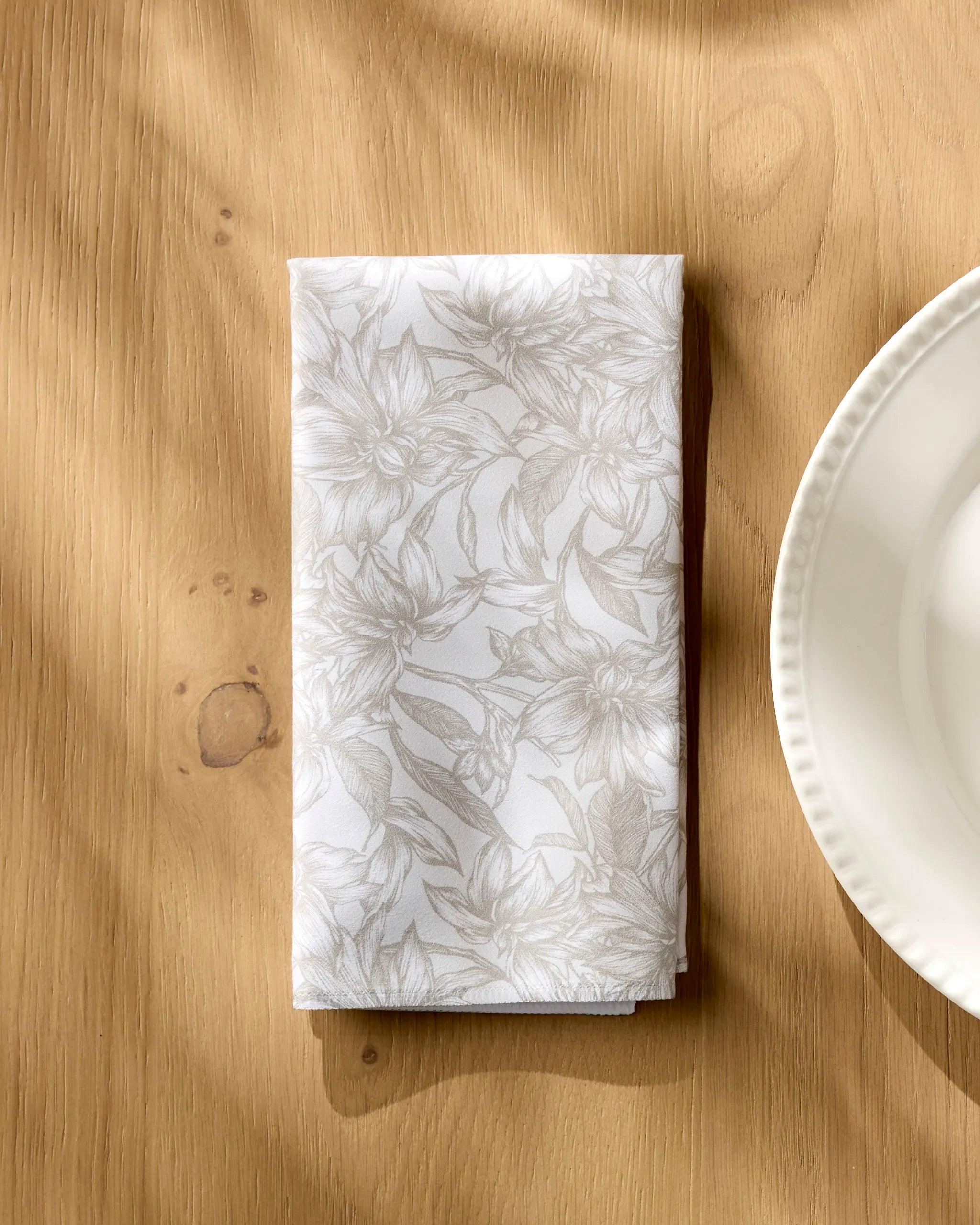 A folded Alba napkin by Geometry, featuring a subtle floral pattern, rests on a light wooden table beside a white scalloped plate as sunlight casts gentle shadows across this elegant dinner napkin set.