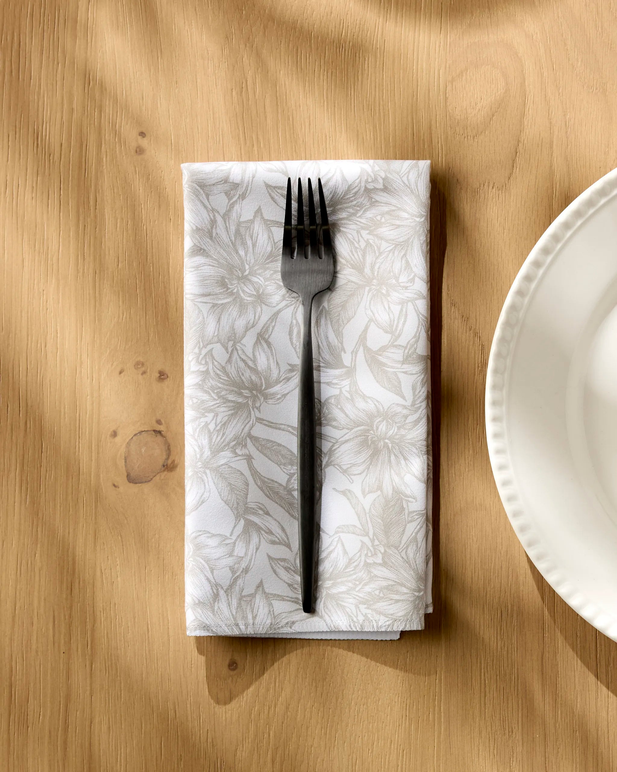 A black fork rests on a folded Alba napkin by Geometry—an absorbent, floral-patterned dinner napkin—on a wooden table next to a white plate, with sunlight casting soft shadows across the scene.
