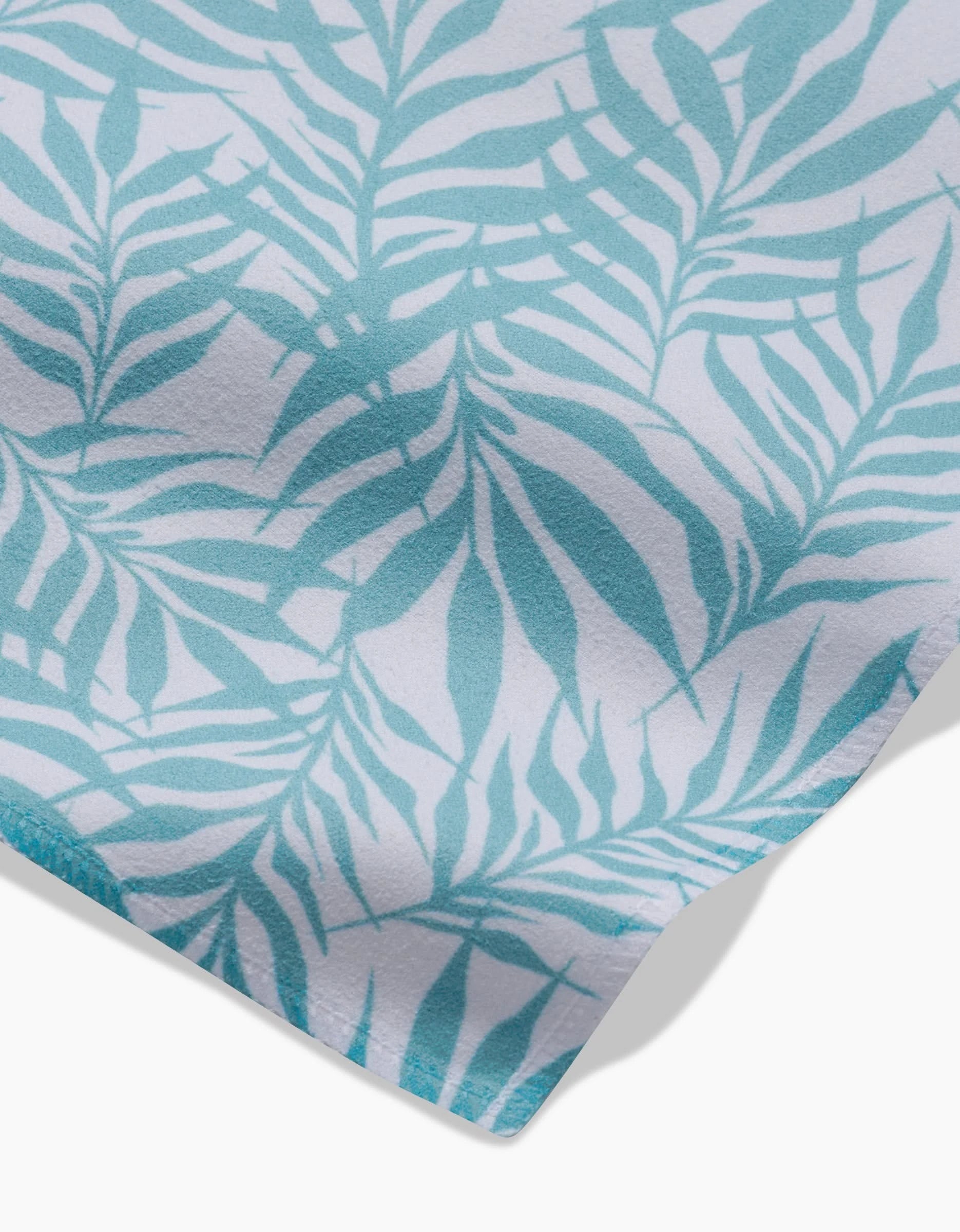 A close-up of the GEOMETRY Aqua Branches napkin shows blue and white tropical, fern-like leaves on a light background, with the edge of this recycled dinner napkin visible in the bottom right corner.