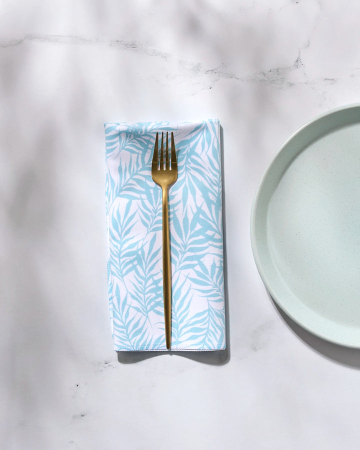 A gold fork rests on a folded, white washable GEOMETRY "Aqua Branches" napkin with a light blue leaf pattern, next to a light blue plate on a white marble surface.
