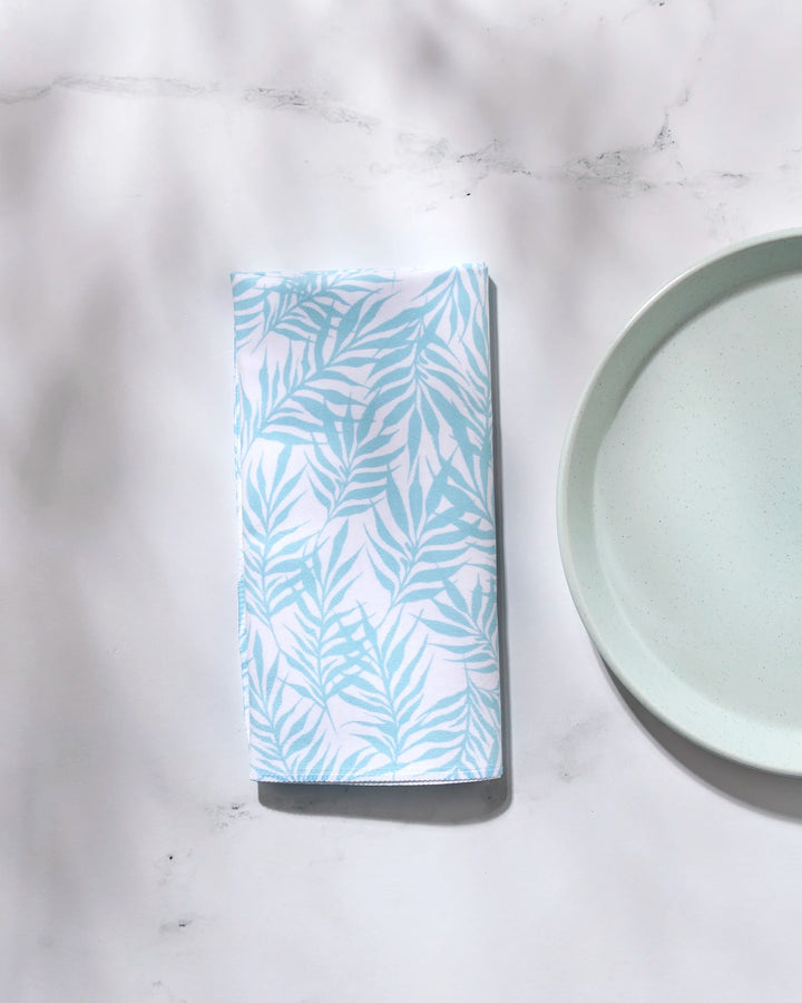 The GEOMETRY Aqua Branches washable cloth napkin with a light blue leaf pattern is folded beside a pale blue plate on a white marble surface with soft shadows.