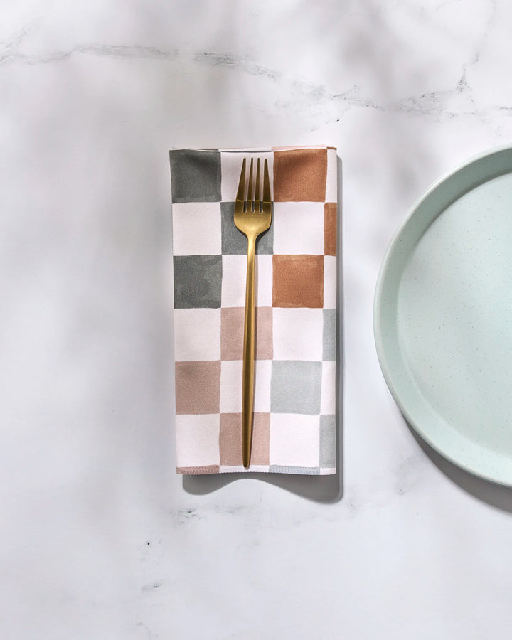 A gold fork rests on the folded GEOMETRY Autumn Checkers reusable napkin, featuring gray, tan, and white squares, placed beside a light blue plate on a white marble surface.