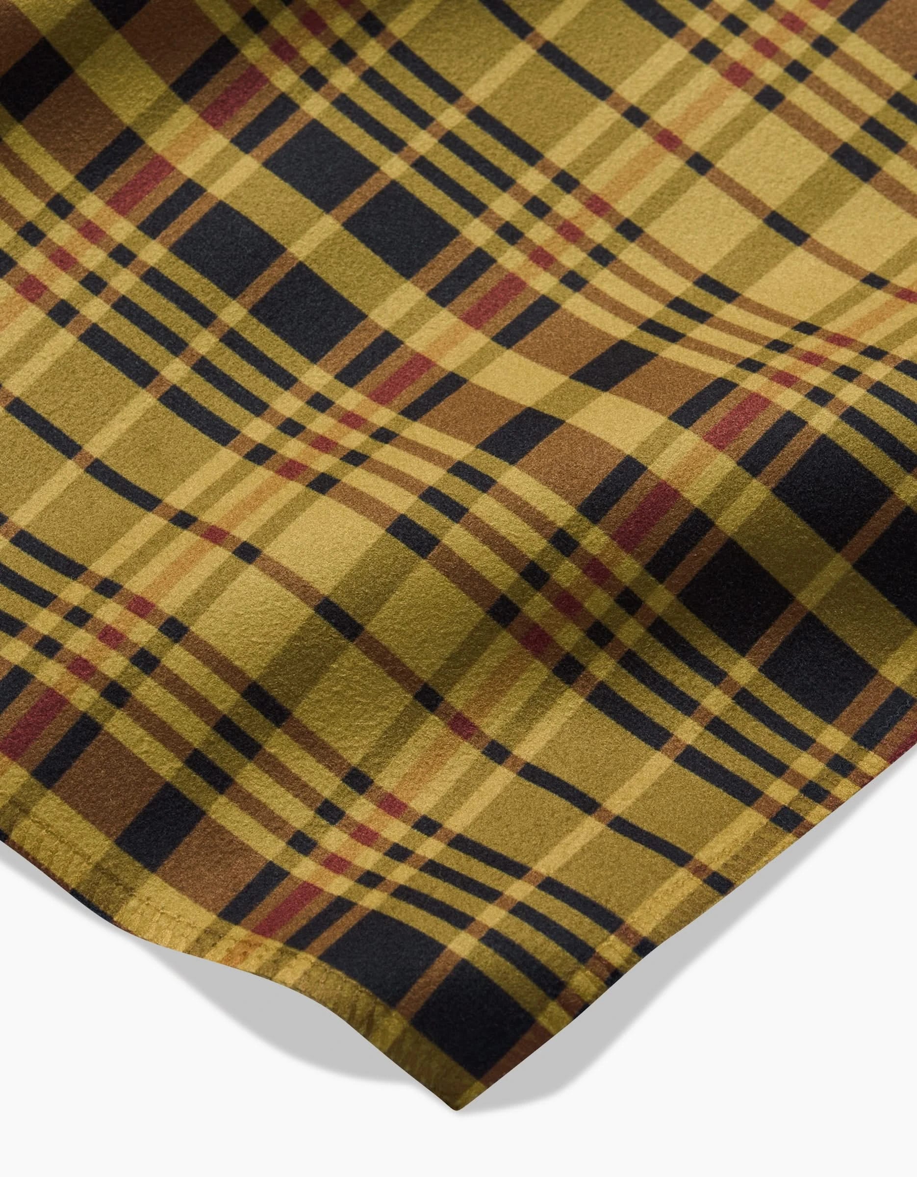 A close-up of the Danelys Sidron Autumn Plaid napkin shows its soft, textured yellow, brown, black, and red fabric laid flat with a slight fold casting a small shadow on a white background.