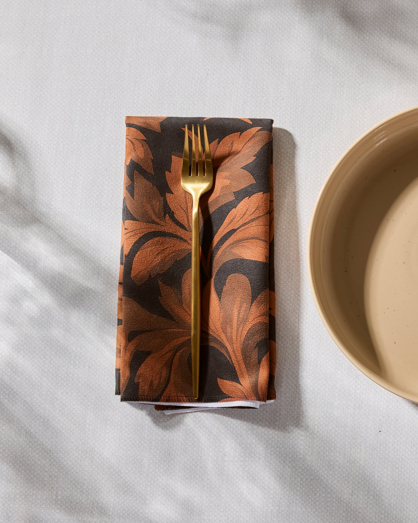 A gold fork rests on Geometry’s Autumnal Classic Foliage absorbent dinner napkin, featuring an orange leaf pattern, atop a white textured tablecloth beside a beige plate.