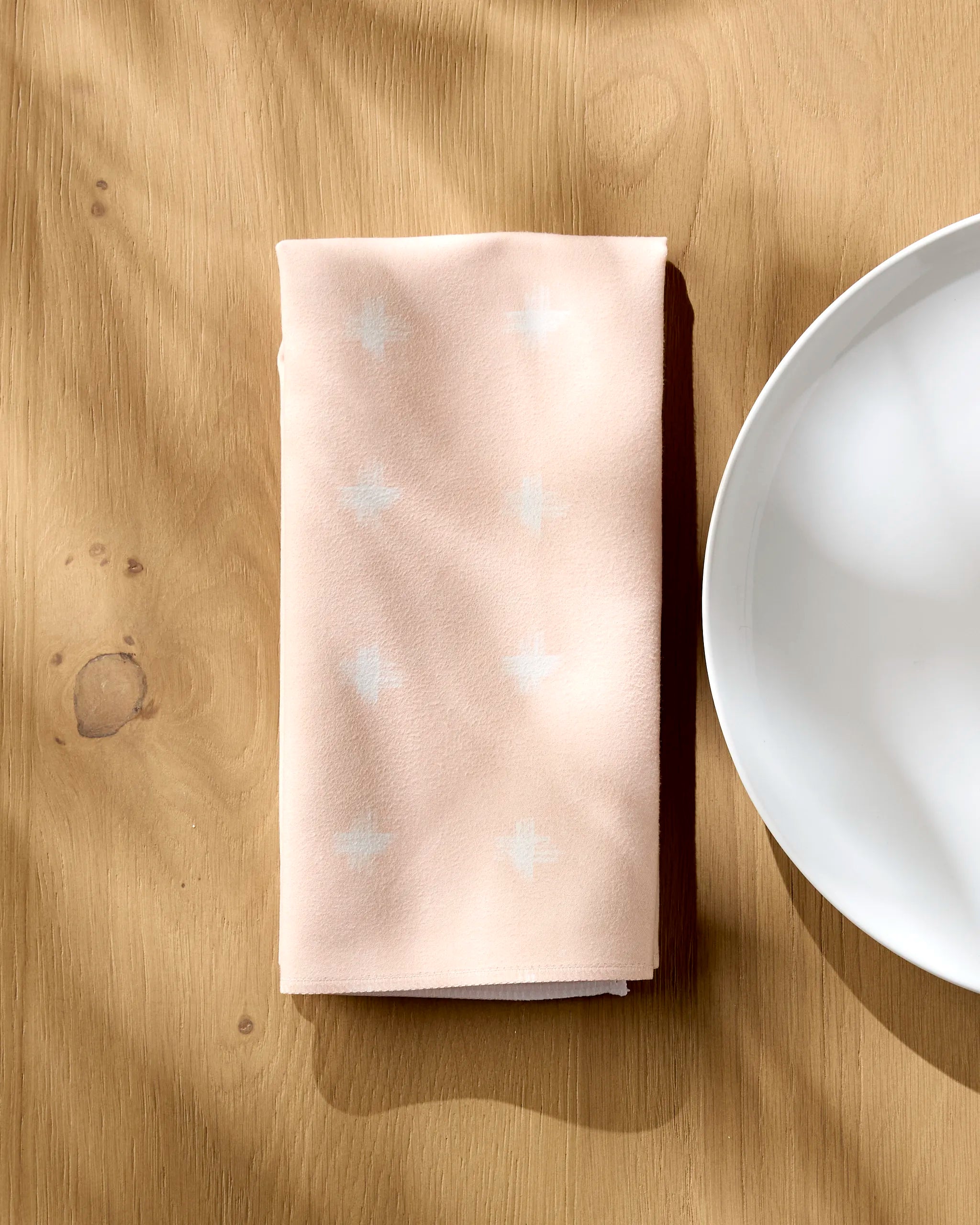 A folded pale pink Aveline Geosuede Fabric napkin by Amy MacCready, with subtle white cross patterns, rests beside a white plate on light wood. Sunlight casts gentle shadows—an elegant addition to any dinner napkin set.